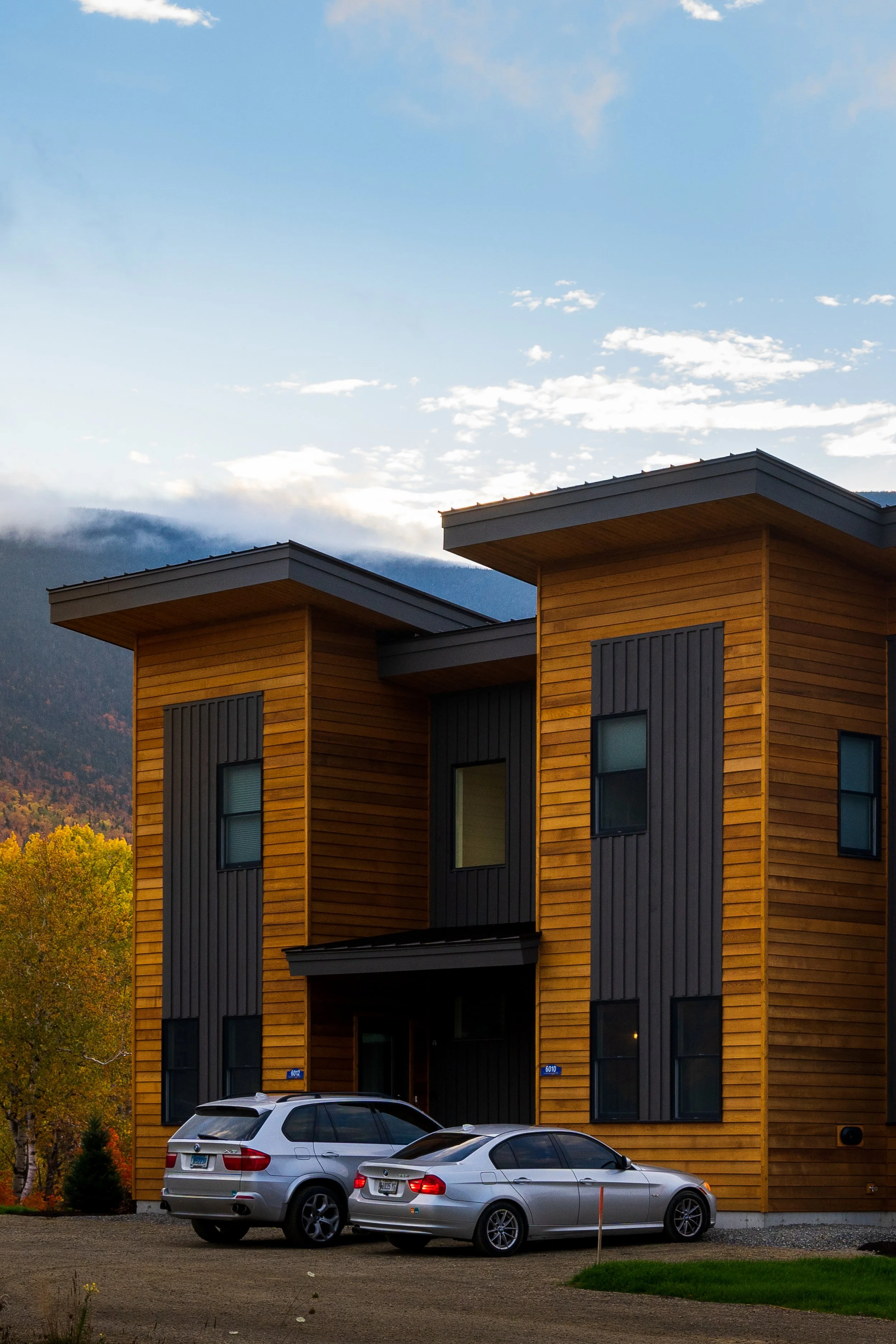 Modern apartment building with wooden and dark panel exterior, three stories, located in a mountainous area with trees showing autumn foliage, and two cars parked in front.