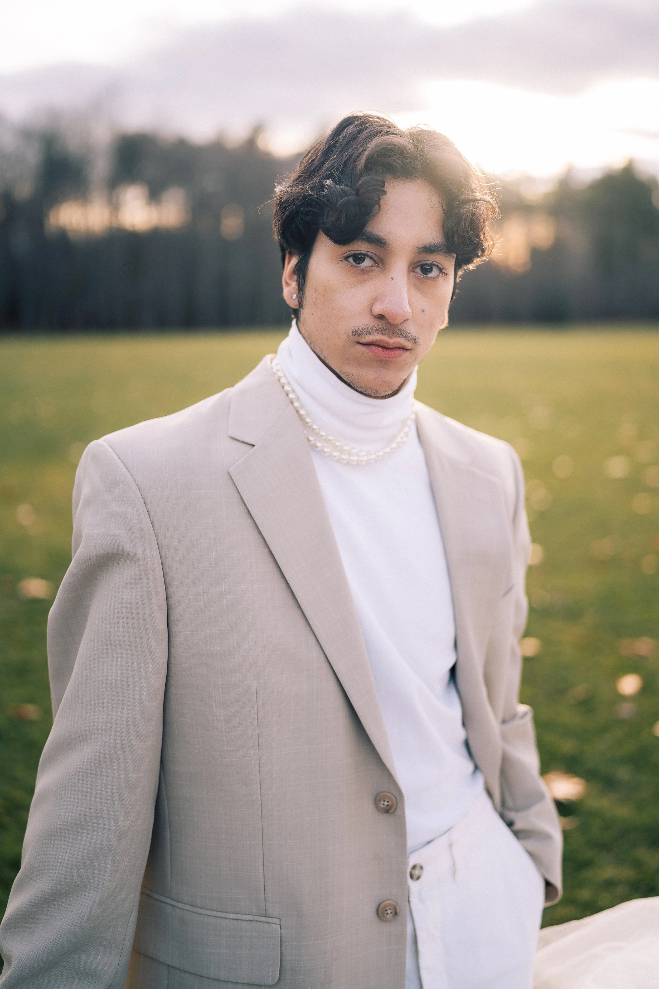 A young person in a beige suit and white turtleneck with pearl necklace stands outdoors on a grassy field at sunset, with trees in the background.