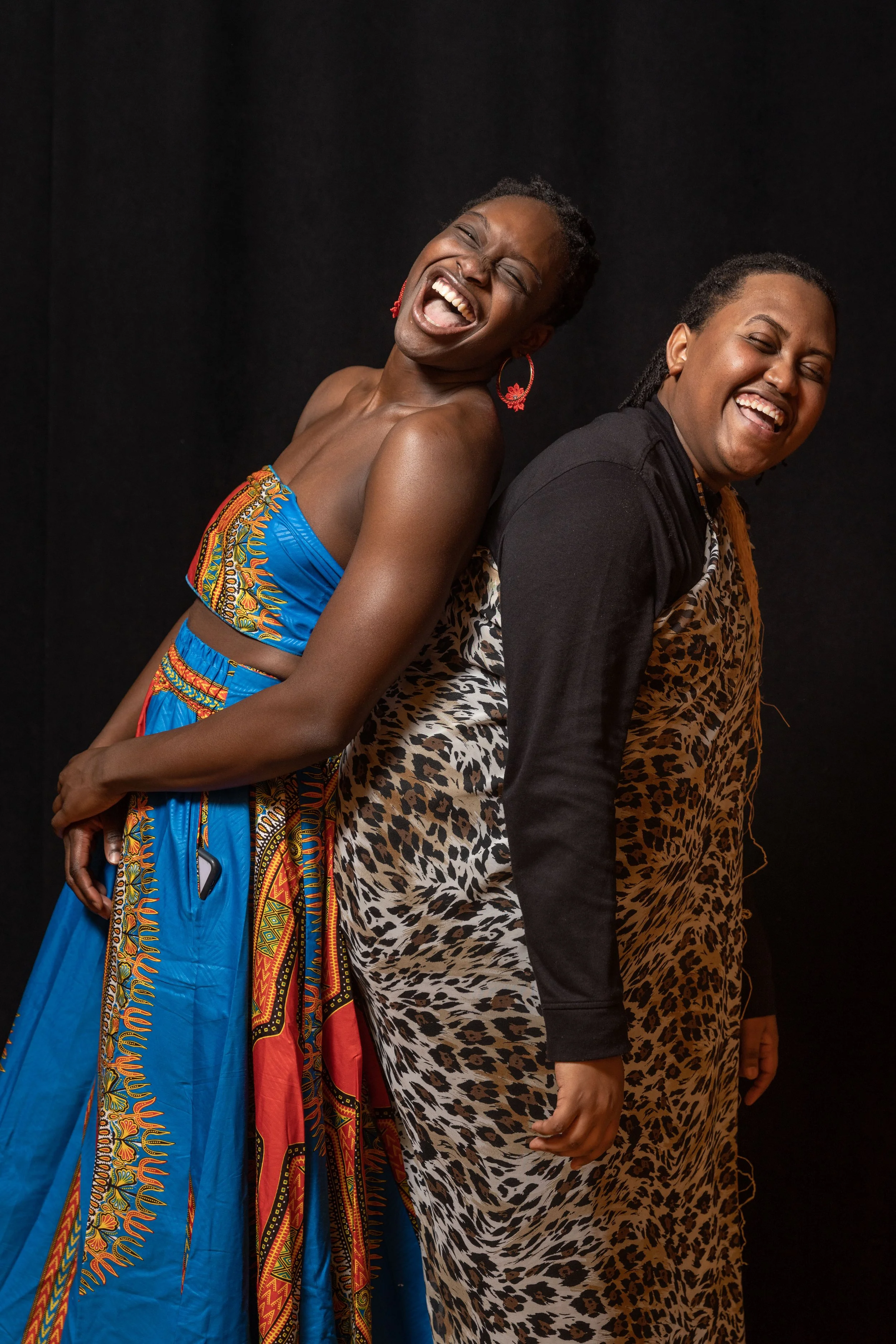 Two women standing back to back, laughing. One is wearing a colorful traditional dress, and the other is in a leopard print dress and black jacket, against a black background.