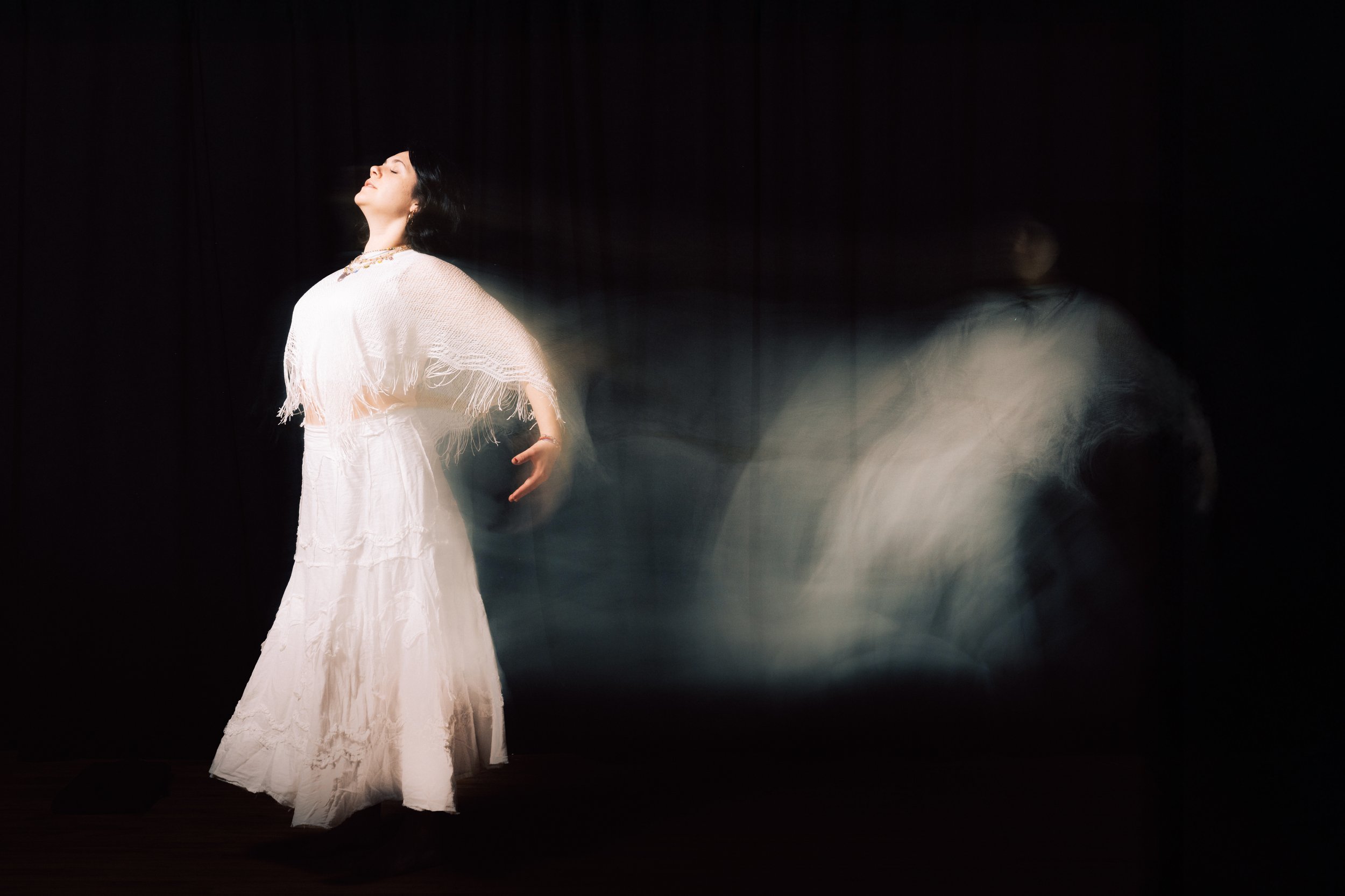 A woman in a flowing white dress and shawl stands with her eyes closed and head tilted back in a dark setting. Light trail effects create a ghostly impression around her.