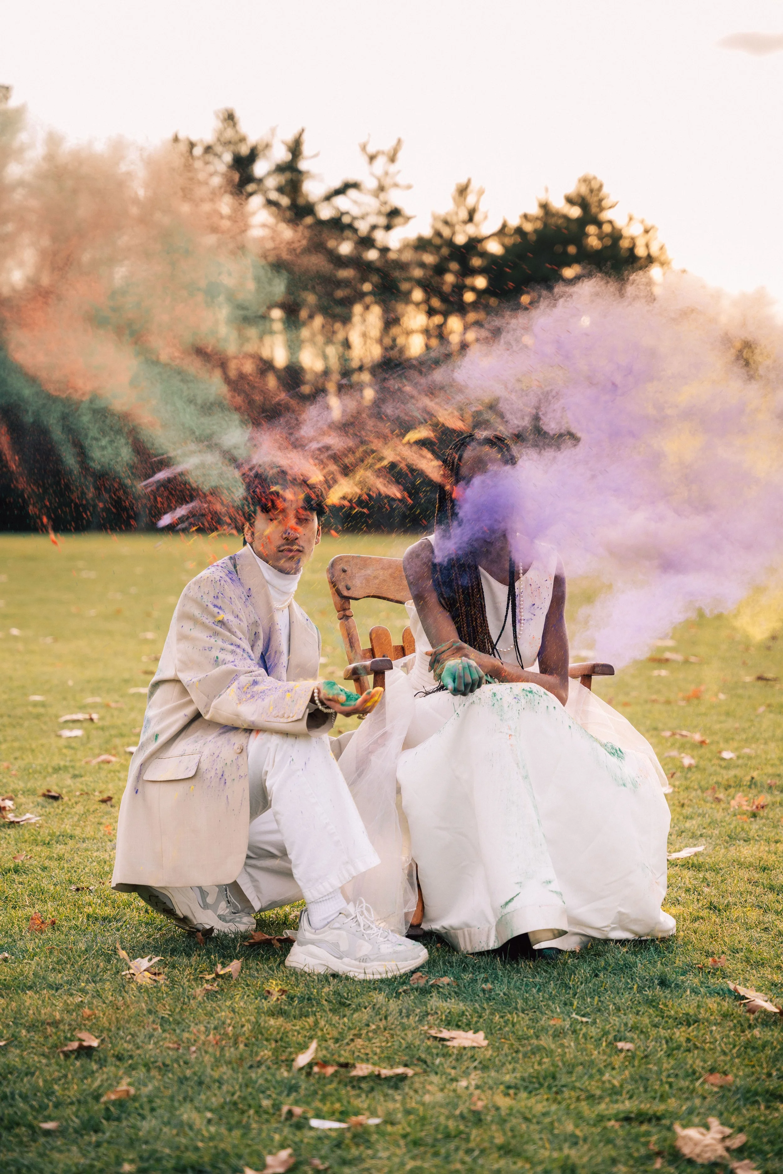 Two people wearing white outfits sitting outdoors on a grassy field with trees in the background. They are releasing colorful smoke and powder.