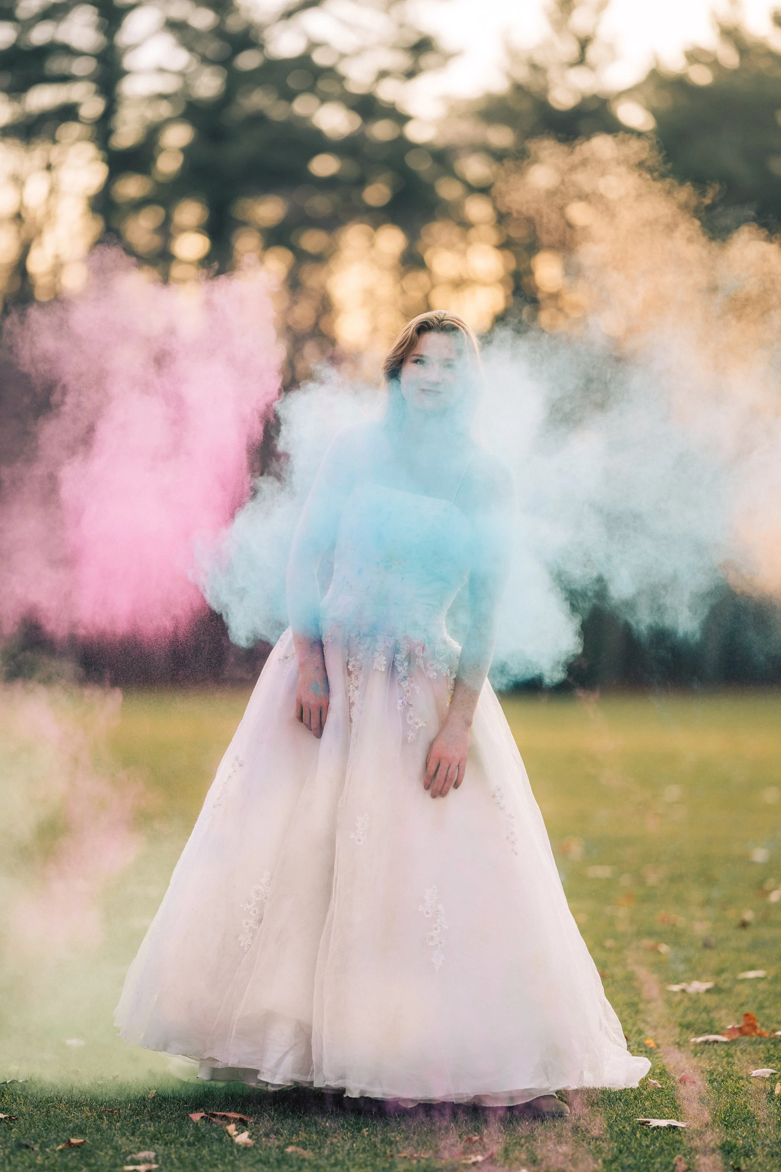 A woman in a white dress standing outdoors on grass, surrounded by colorful smoke clouds in pink, blue, and orange with trees in the background during sunset.