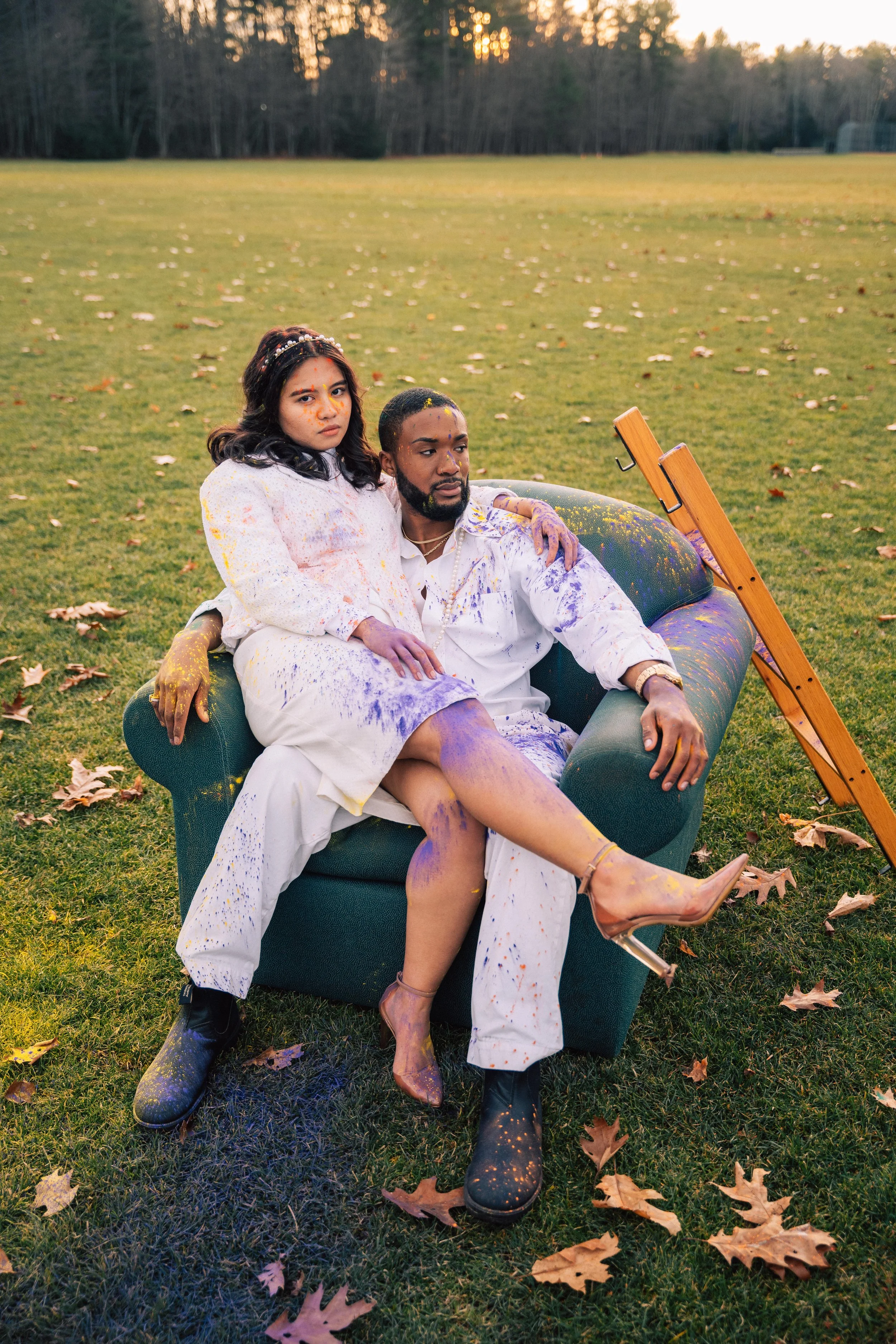 A man and woman sitting on a green couch outdoors on a grassy field with fallen leaves. Both are covered in colorful paint splatters, with the woman sitting on the man's lap and the man wearing boots and the woman wearing high heels. A wooden easel leans on the arm of the couch.