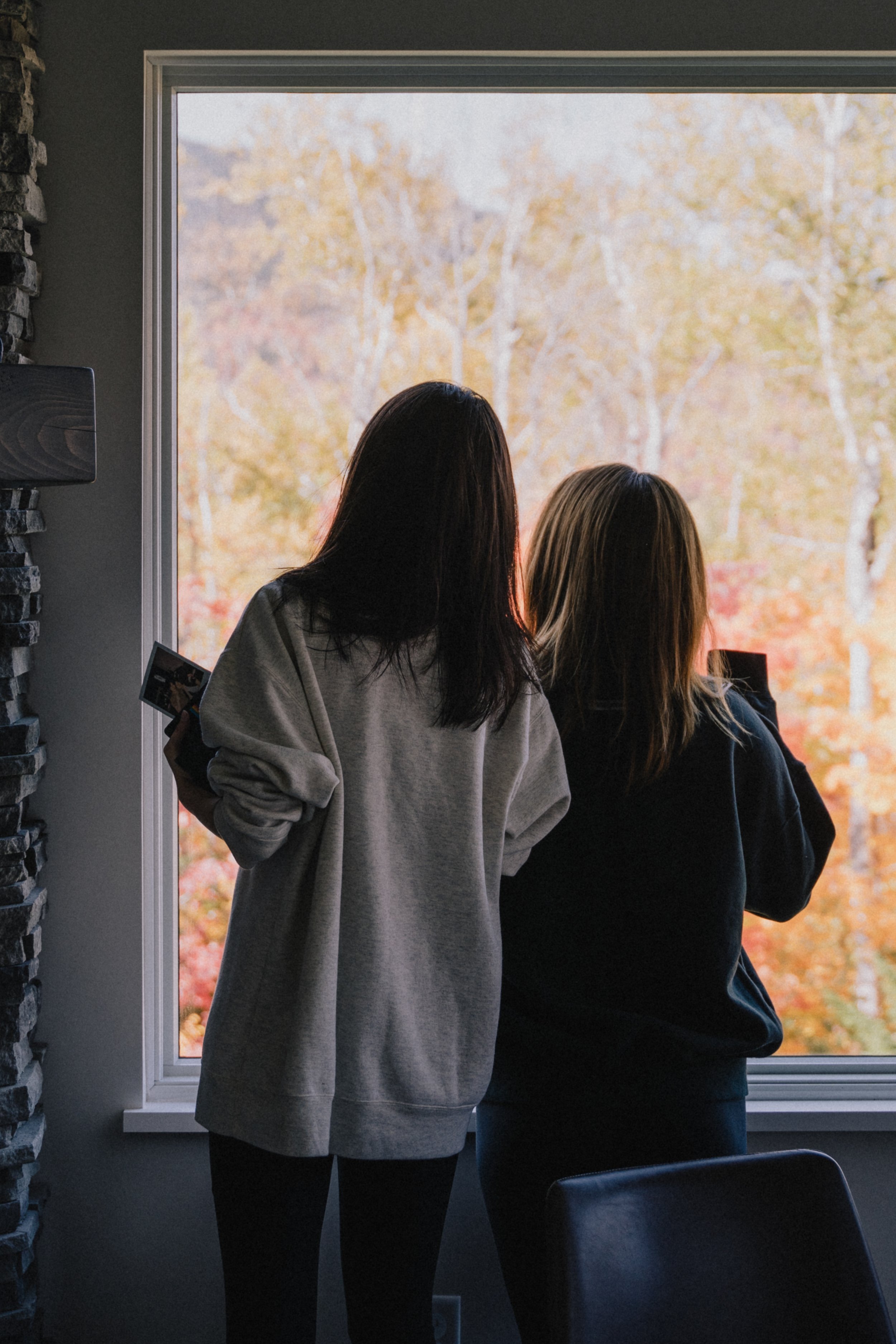 Two women stand by a large window with fall foliage outside, one holding a photo and the other looking out.