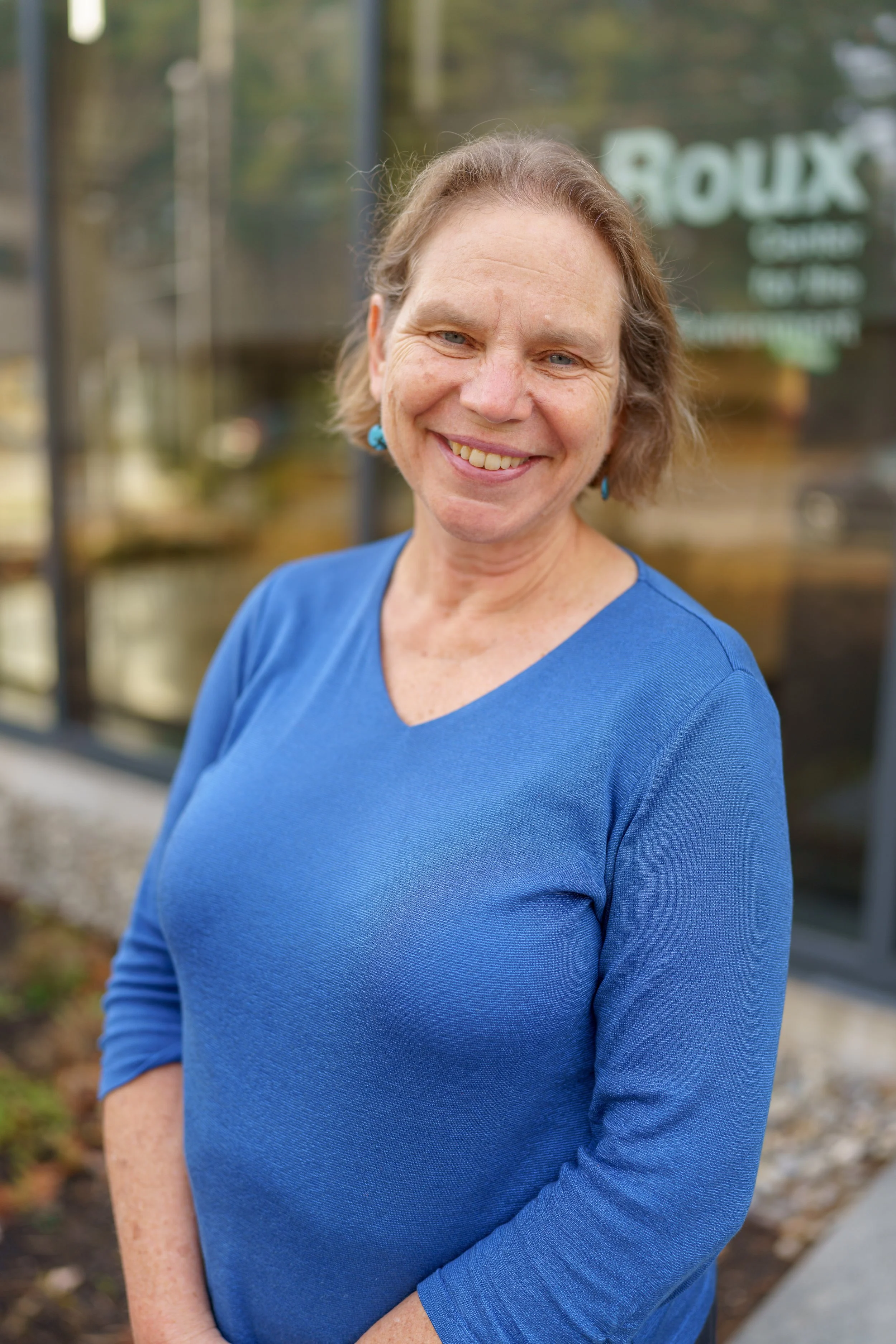 Smiling woman wearing a blue top standing outdoors in front of a glass building with a sign that says 'ROUX'.