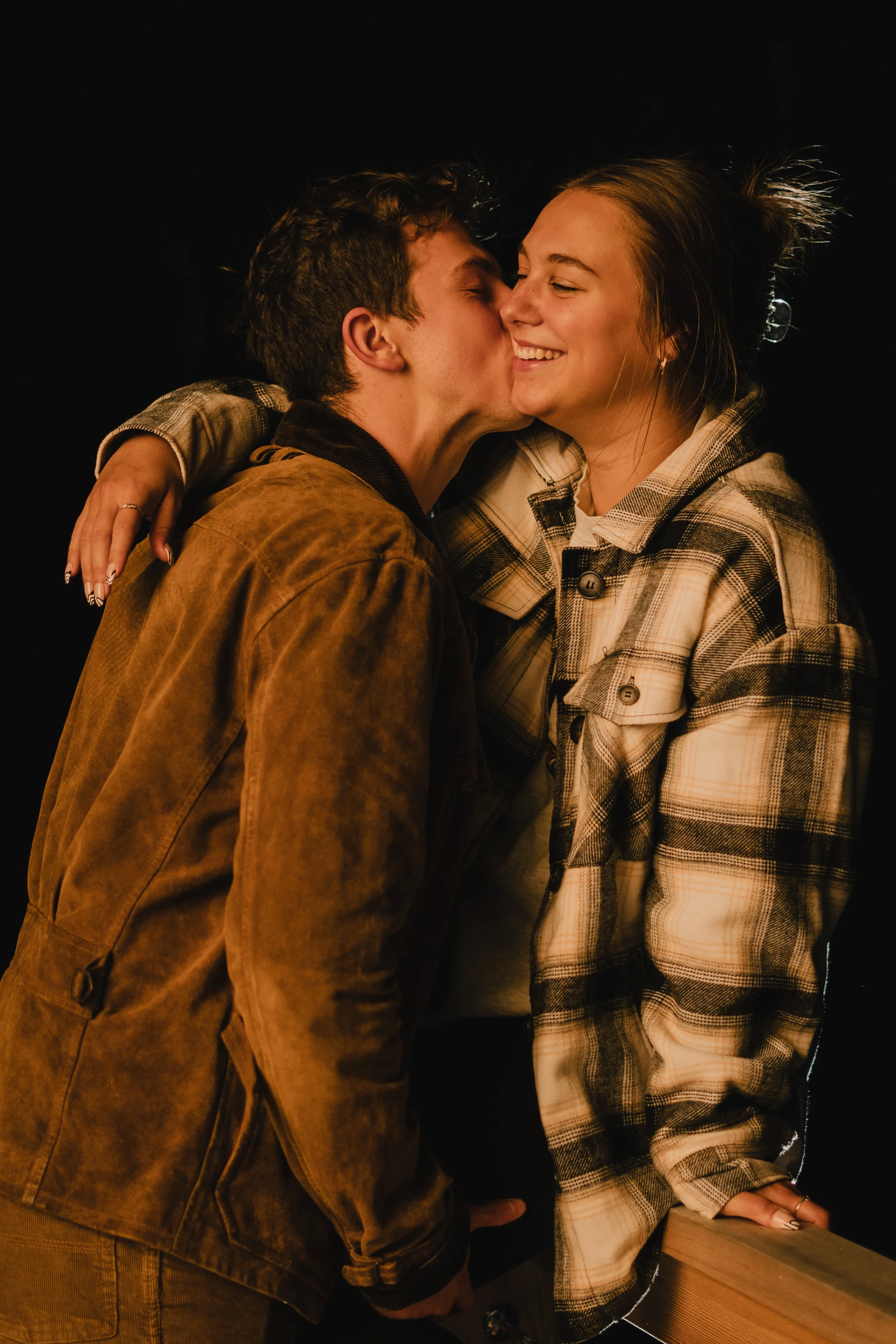 A young couple sharing a kiss, with the man kissing the woman's cheek, both smiling, wearing casual plaid and brown jackets, with a dark background.