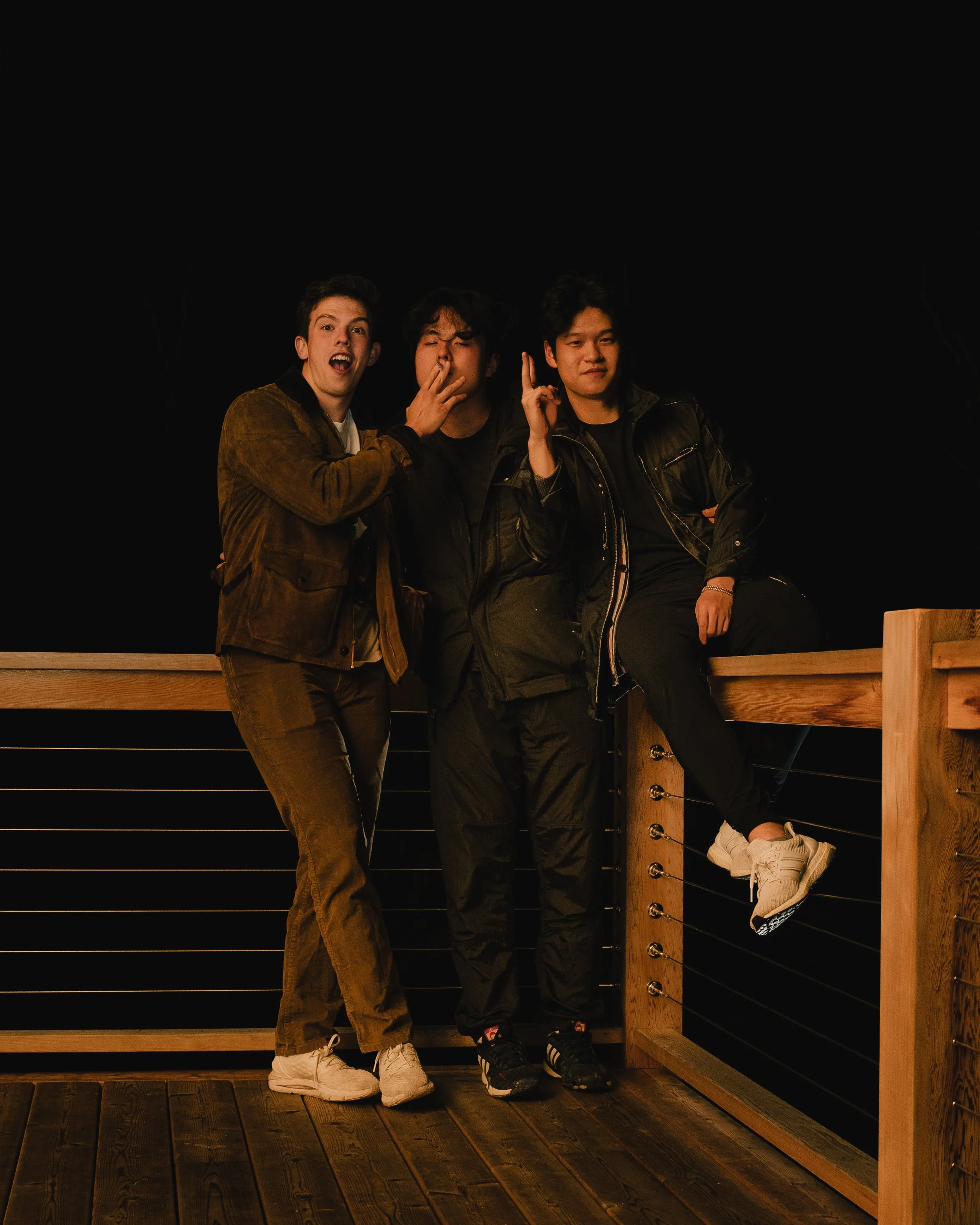 Three young men standing on a wooden deck at night, posing and making gestures.