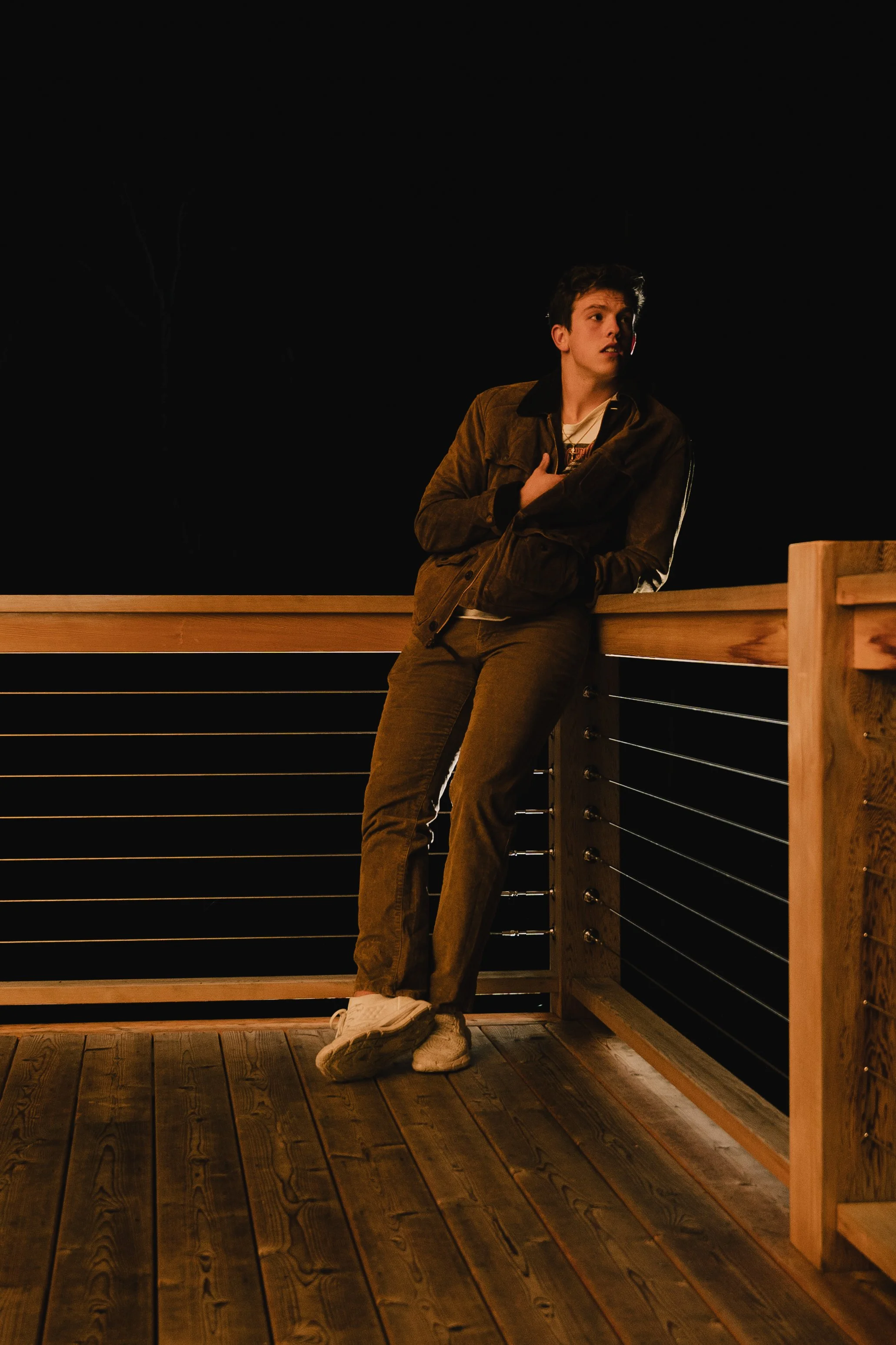 A young man wearing a brown jacket and pants, white sneakers, standing on a wooden outdoor deck at night, leaning against a wooden railing, with a dark background.