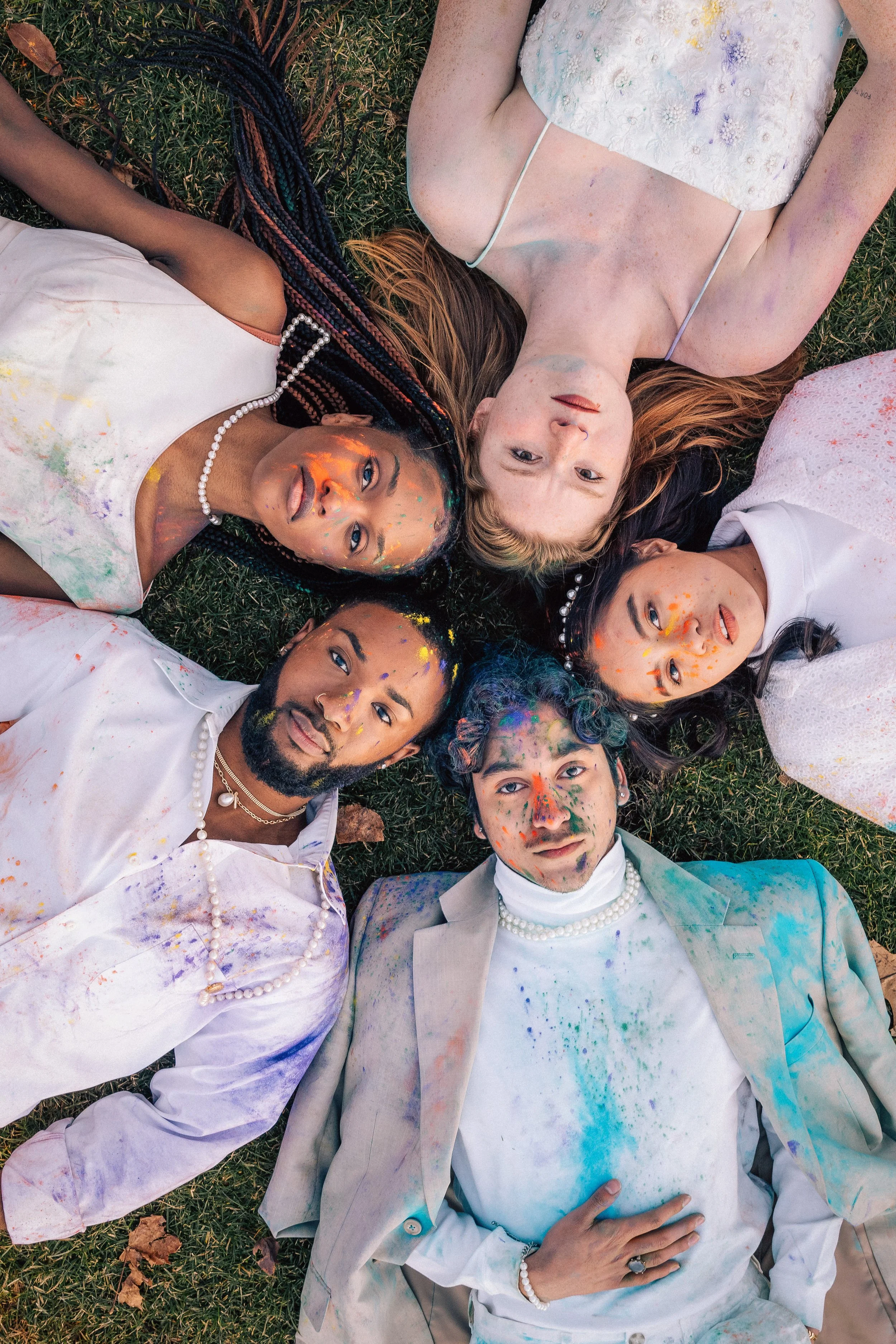Group of six diverse people lying on the grass in a circle, covered in colorful paint splatters, looking up at the camera, dressed in white or light-colored clothing, some wearing pearl jewelry, celebrating a paint or color festival.
