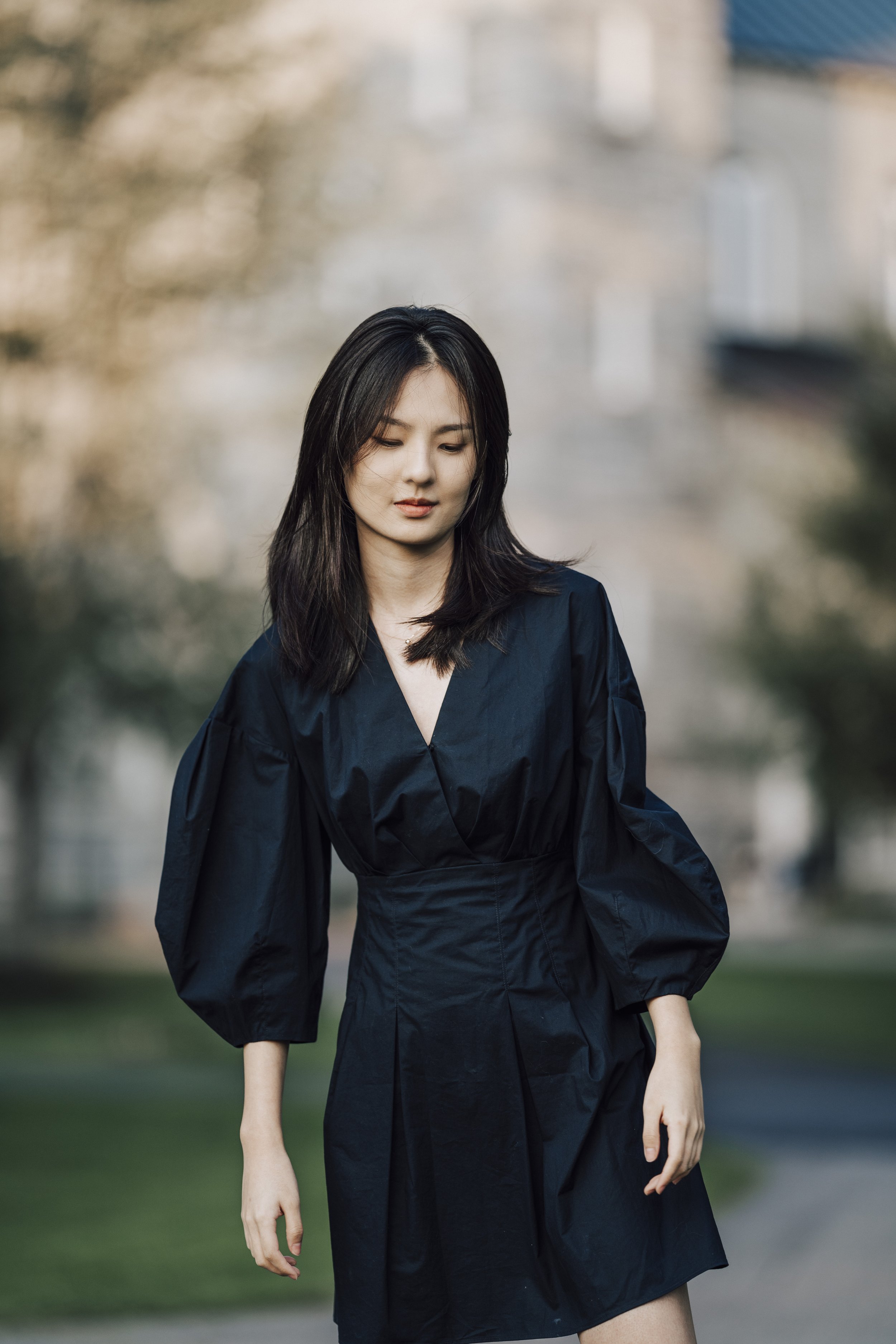 A woman with dark hair wearing a black dress with puffy sleeves, standing outdoors with blurred trees and buildings in the background.