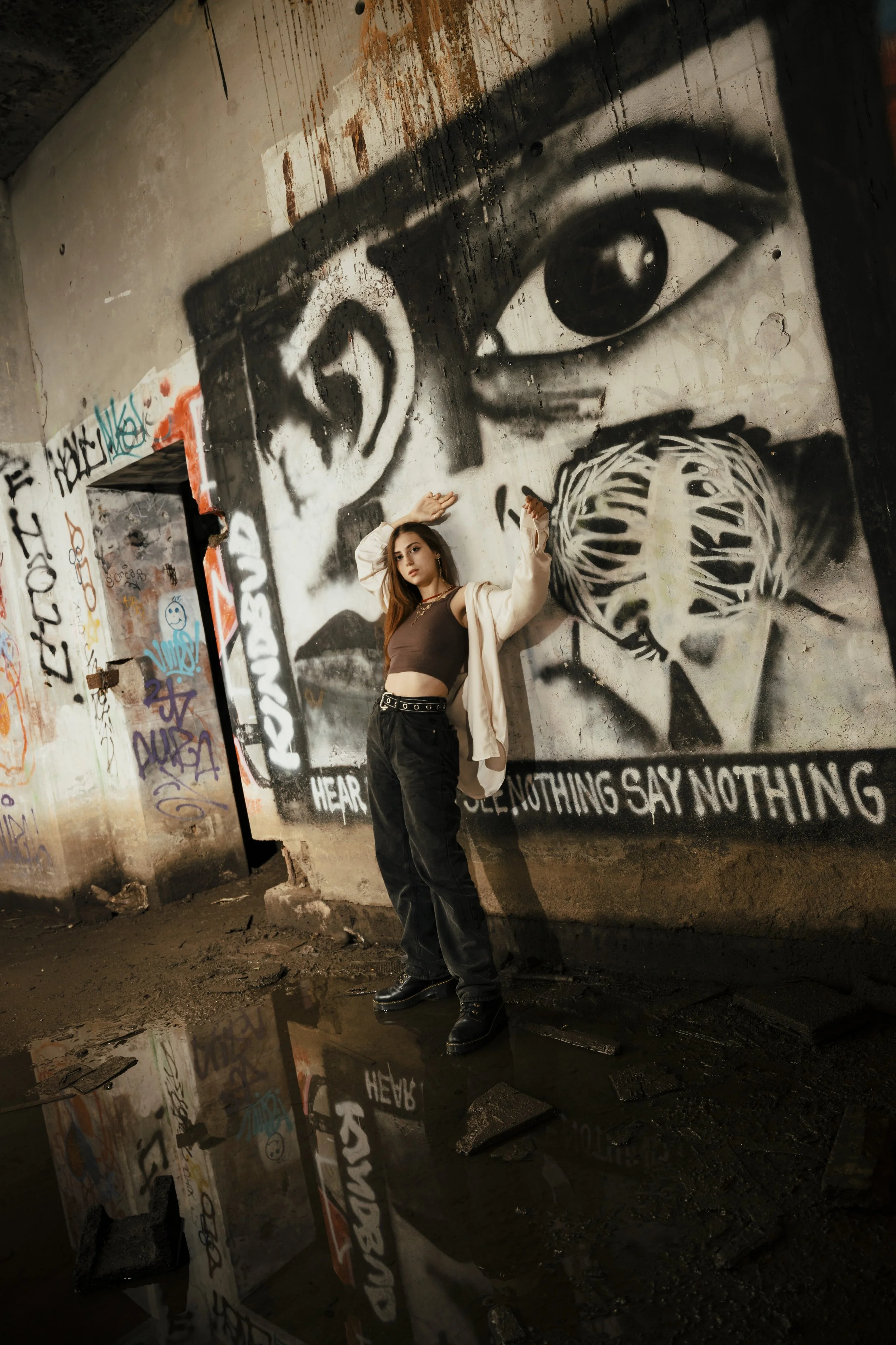 Young woman stands in front of graffiti mural of a large eye and face on a wall in an abandoned, graffiti-covered space, with puddles reflecting the mural on the floor.