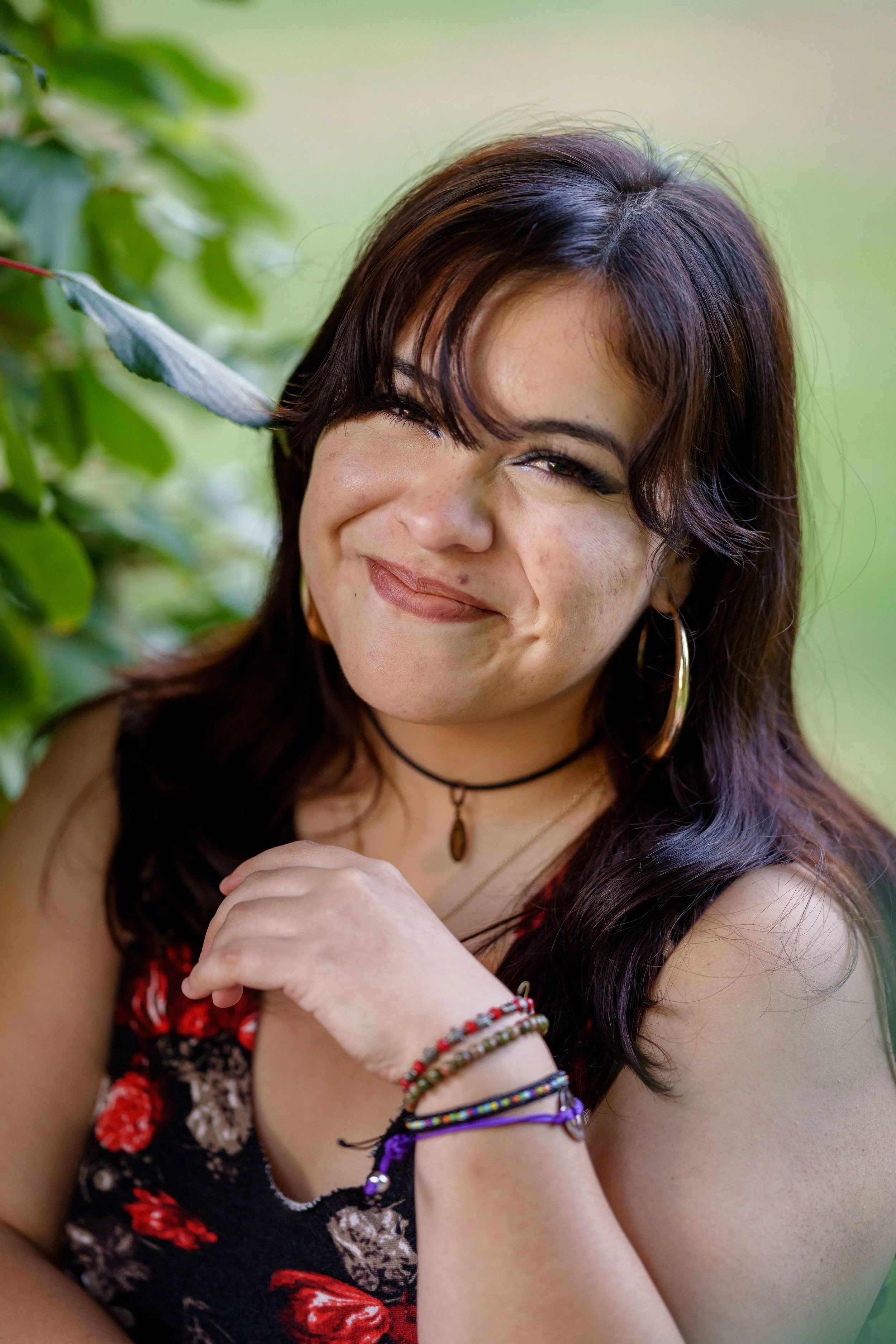A woman outdoors with dark brown hair, winking and smiling, surrounded by green foliage, wearing a floral top, hoop earrings, a choker, and multicolored bracelets.