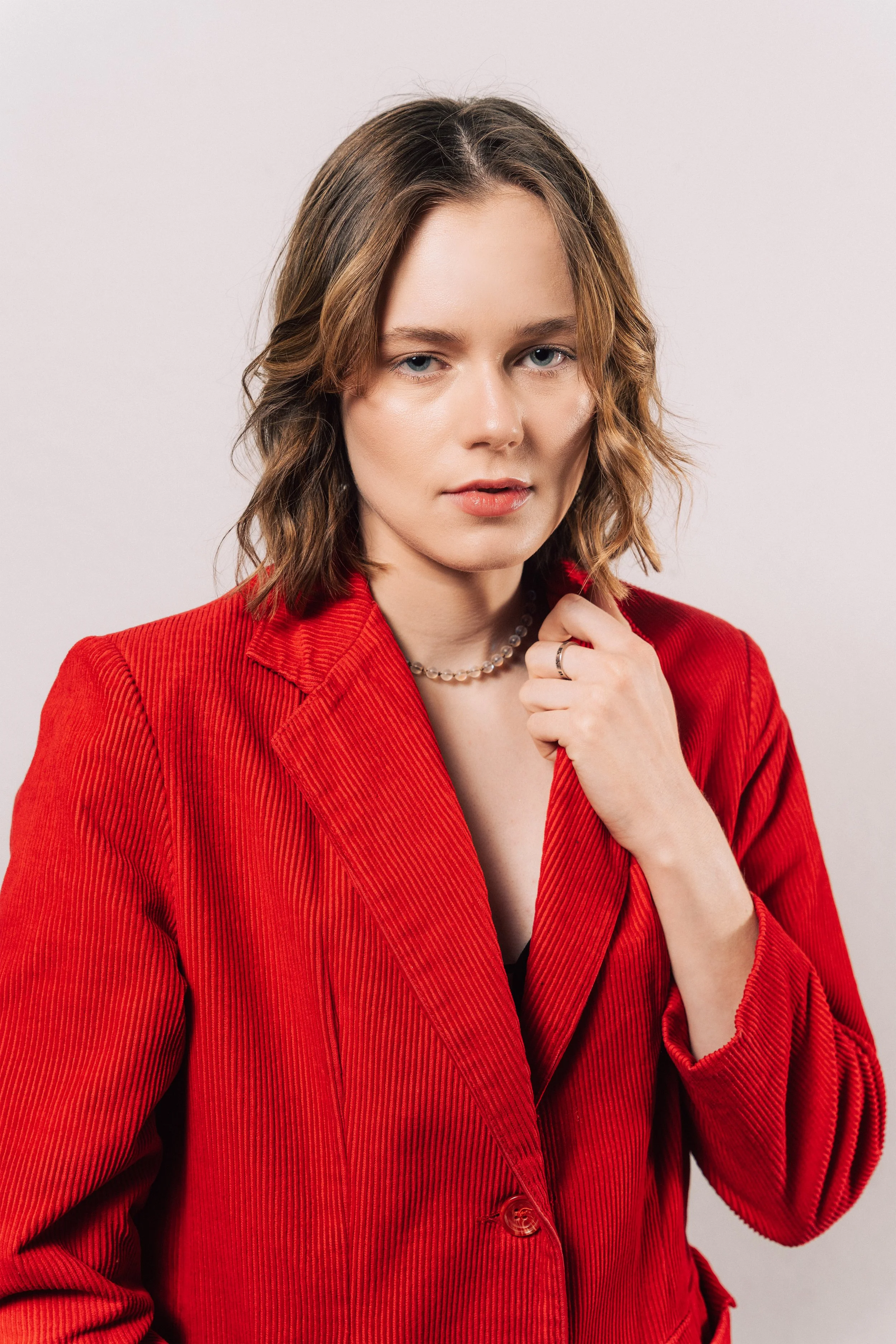 A woman in a red blazer with shoulder-length wavy hair, wearing a pearl necklace, and holding her collar with her right hand.