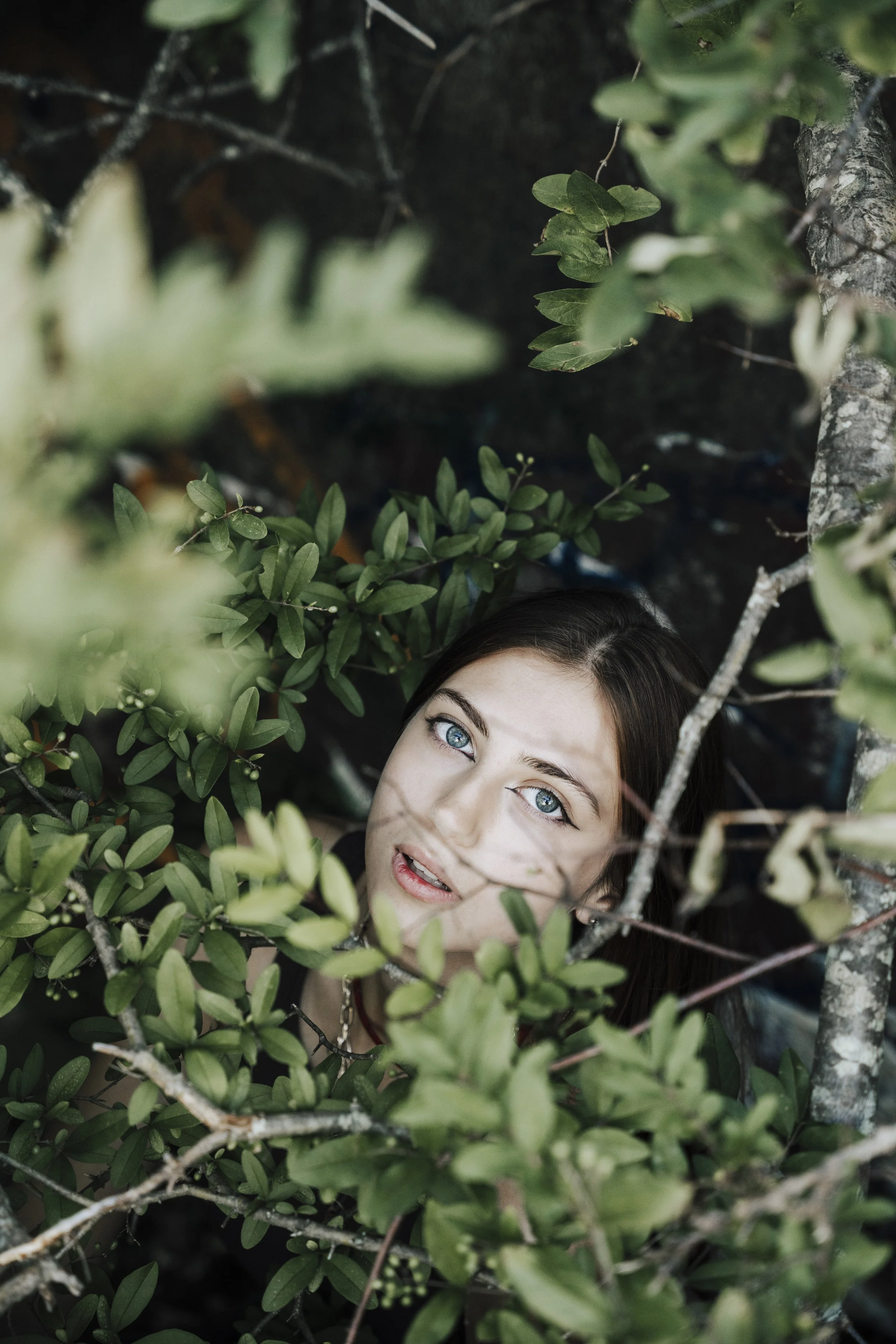 Woman with blue eyes peeking through green bushes.