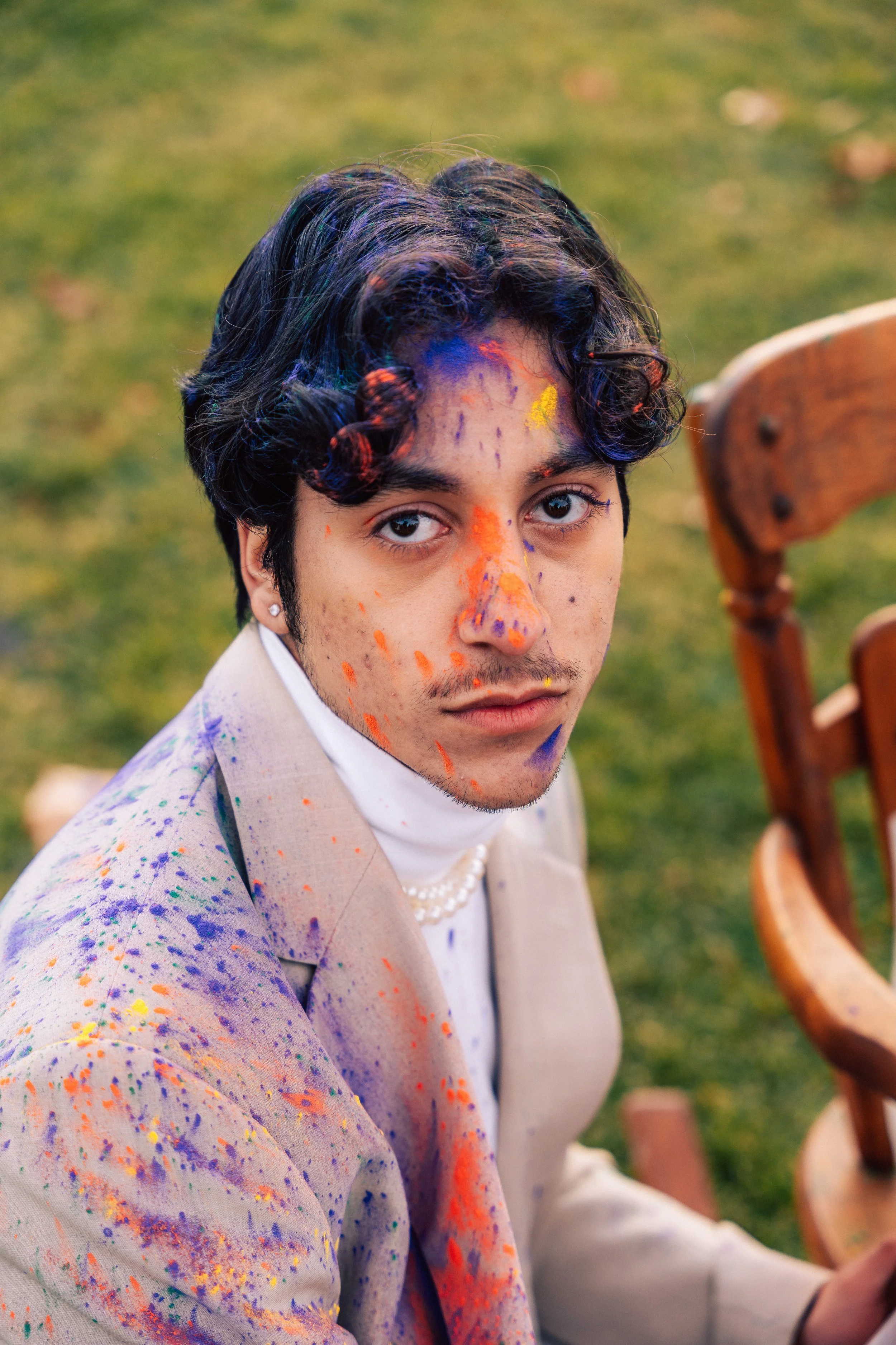 A young man with dark, curly hair and light skin, wearing a beige suit and pearl necklace, covered in colorful paint splatters, sitting outdoors on a grassy area.
