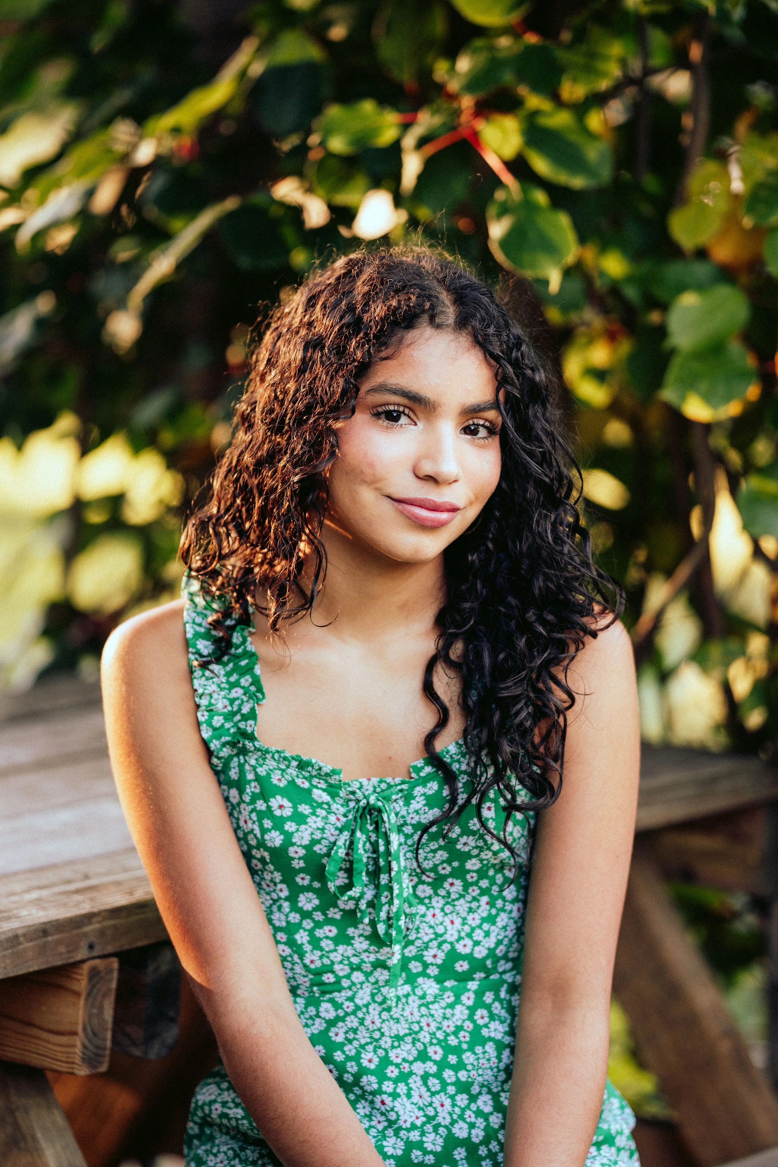 A young woman with dark curly hair is sitting outdoors near a wooden bench with green foliage in the background. She is wearing a green sleeveless dress with white floral patterns and a bow at the neckline, and she is smiling softly.