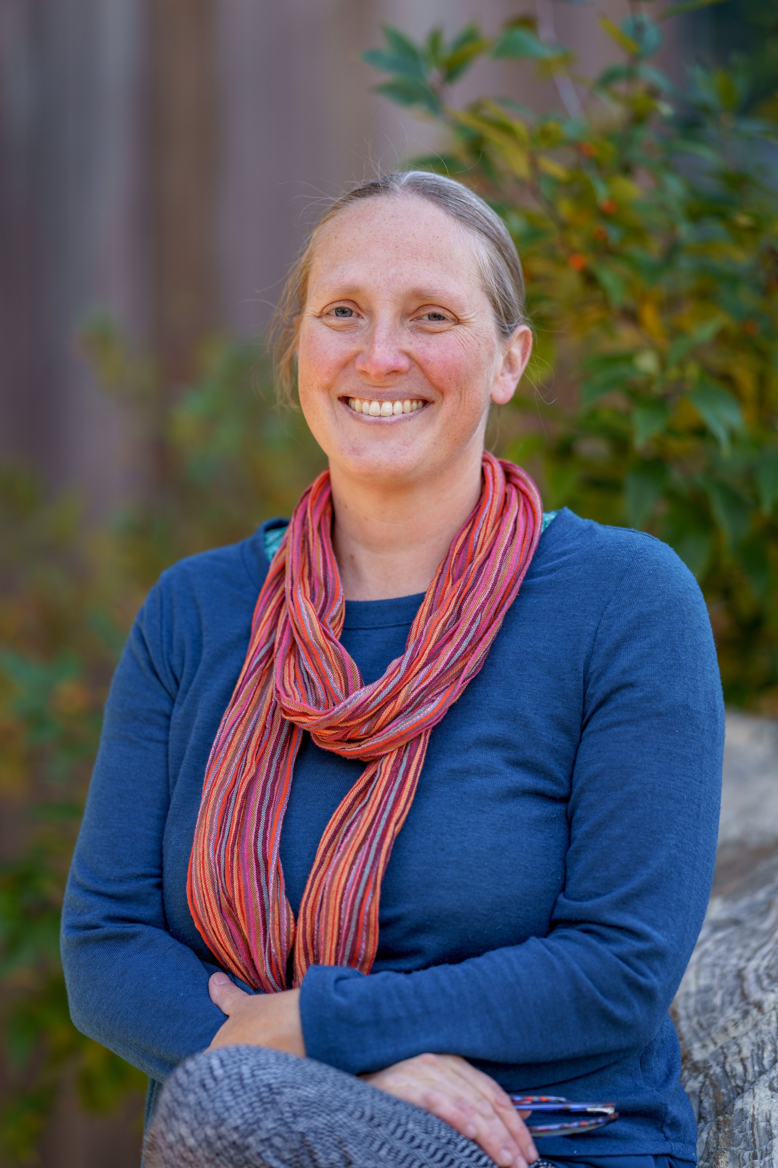 A smiling woman with light skin, wearing a blue long-sleeve shirt and a multicolored scarf, sitting outdoors in front of a blurred background of greenery and trees.