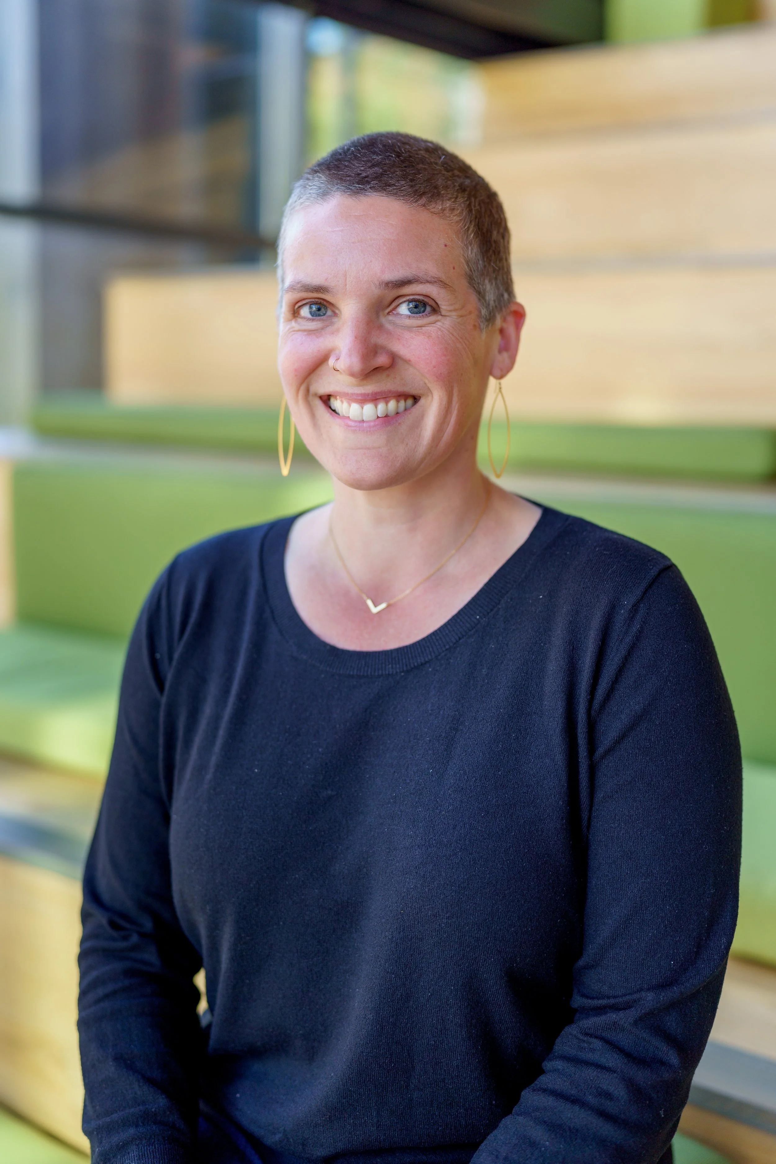 A woman with short hair, wearing a black long-sleeve shirt and gold jewelry, sitting indoors in front of green and yellow seating.