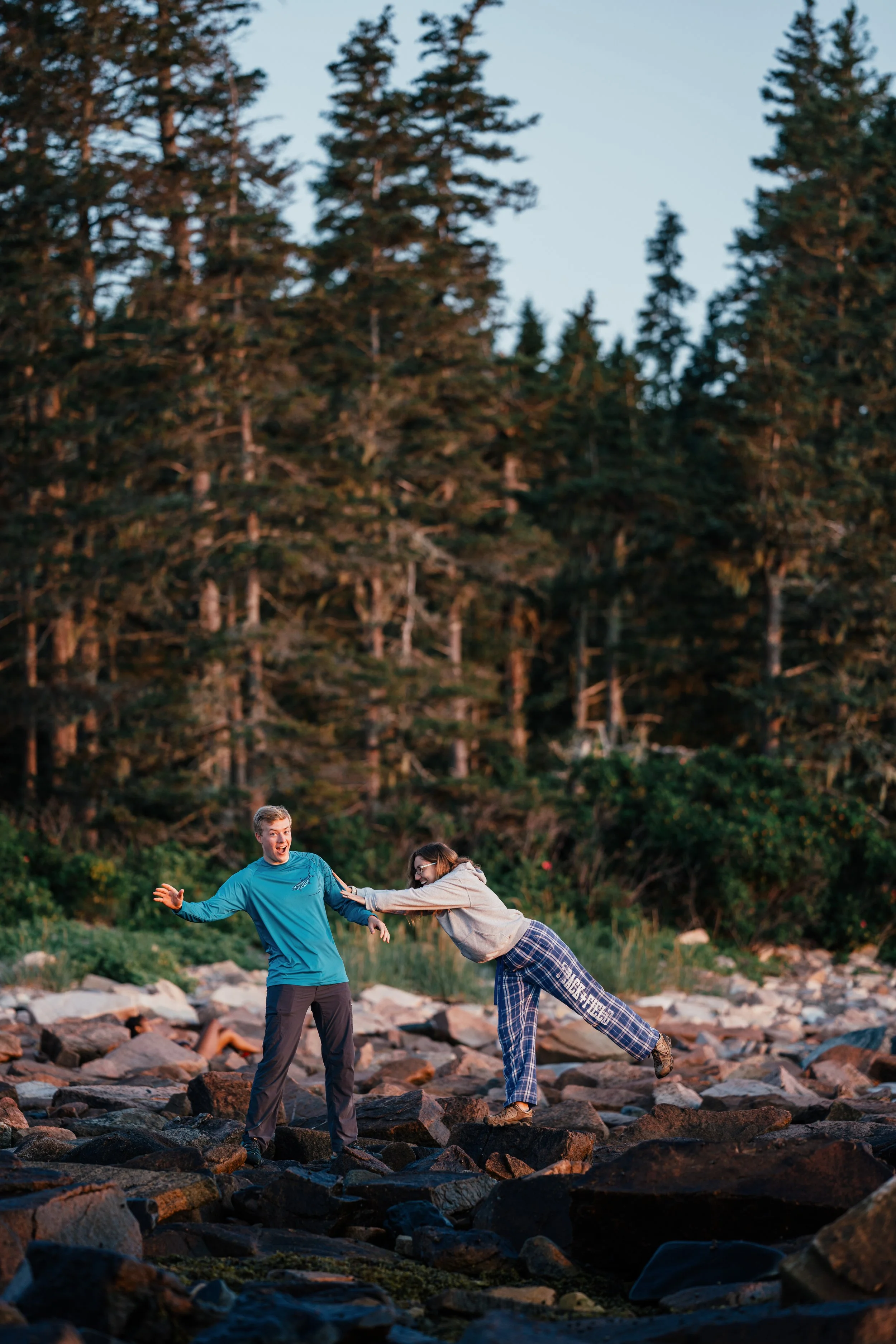 Two people on a rocky outdoor terrain, one man and one woman, playing and laughing with each other during sunset among a forested area.