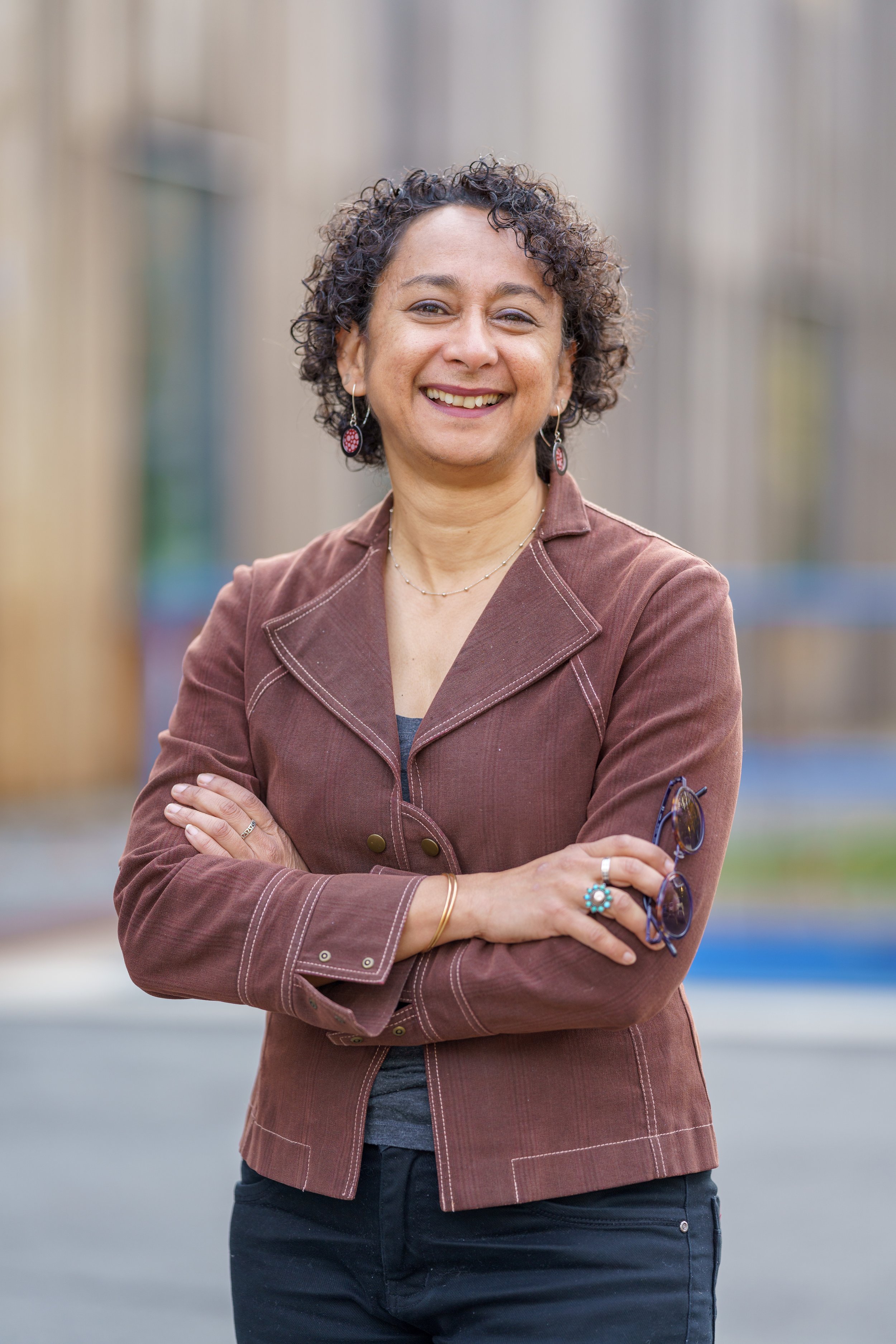 A woman with curly hair smiling and standing outdoors with arms crossed, wearing a brown jacket and earrings.