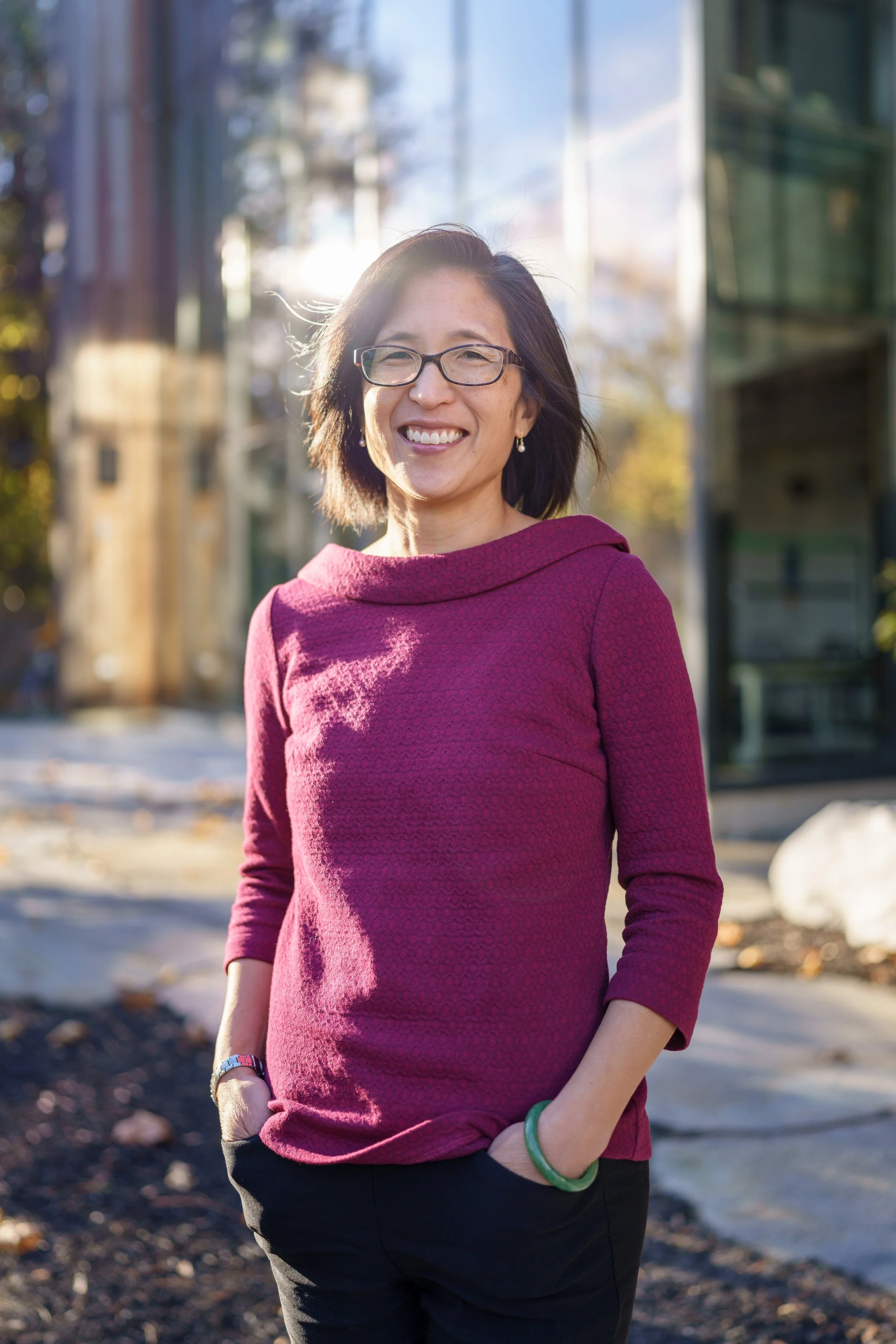 A woman with short dark hair and glasses smiling outdoors in front of a modern glass building on a sunny day, wearing a magenta long sleeve top and black pants.