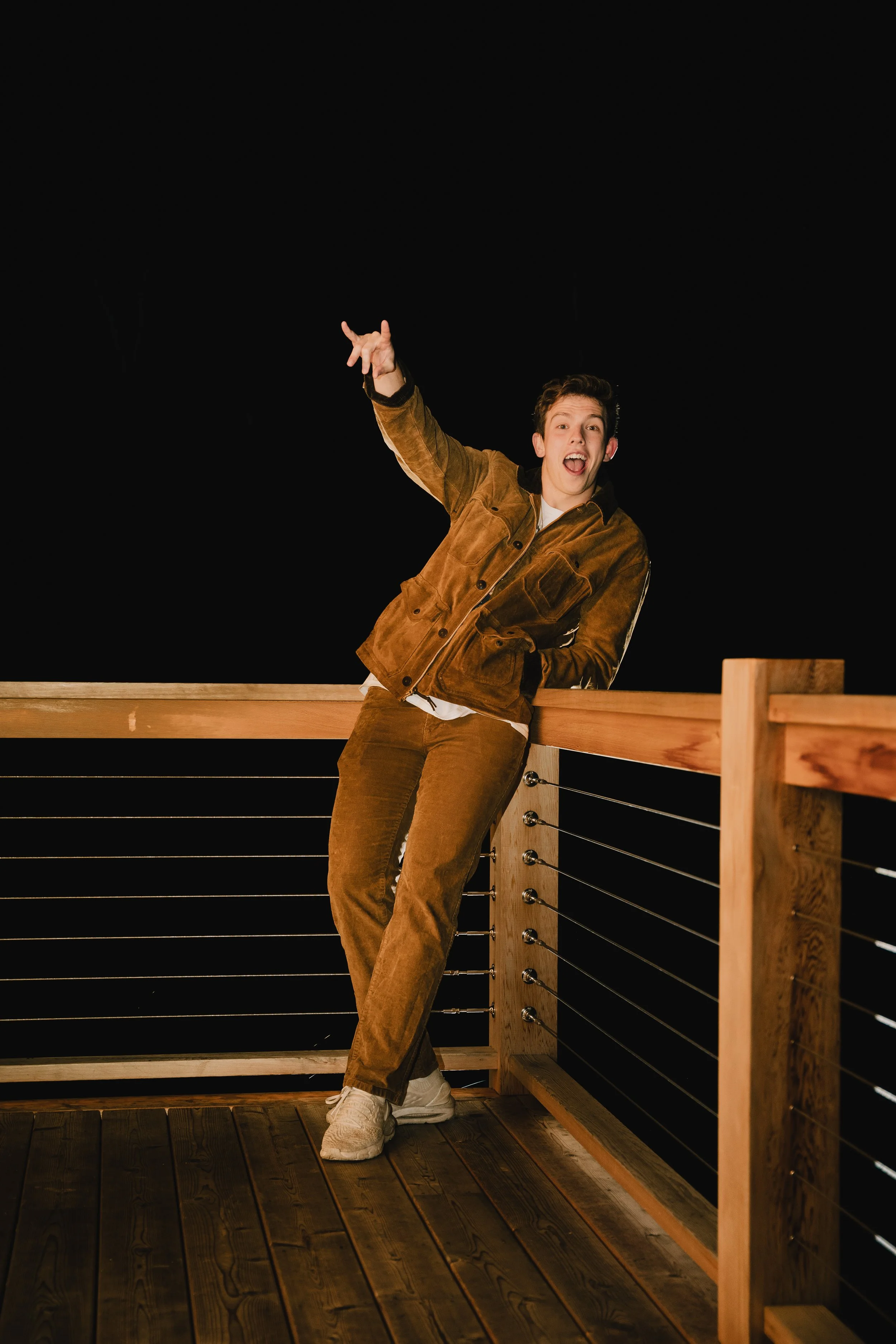 Young man in brown corduroy outfit leaning on a wooden railing, smiling and making a hand gesture against a dark night sky.