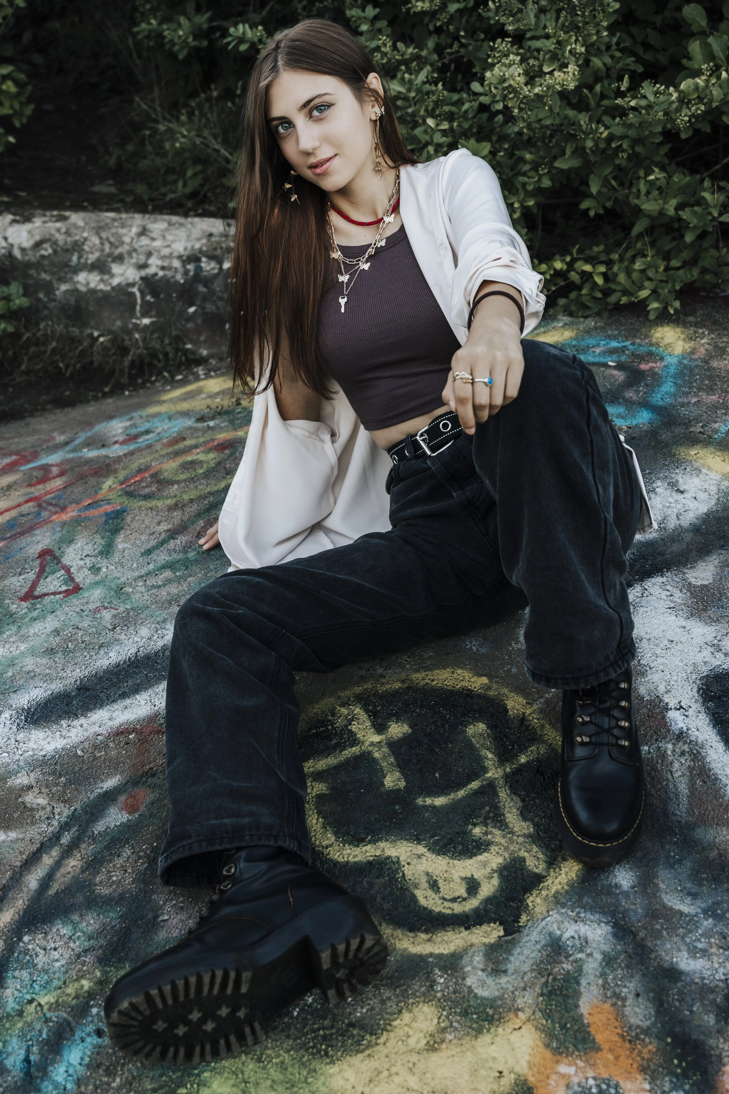 Young woman with long brown hair, blue eyes, wearing a black crop top, white jacket, black jeans, and black combat boots, sitting on a graffitied pavement outside with green bushes in the background.