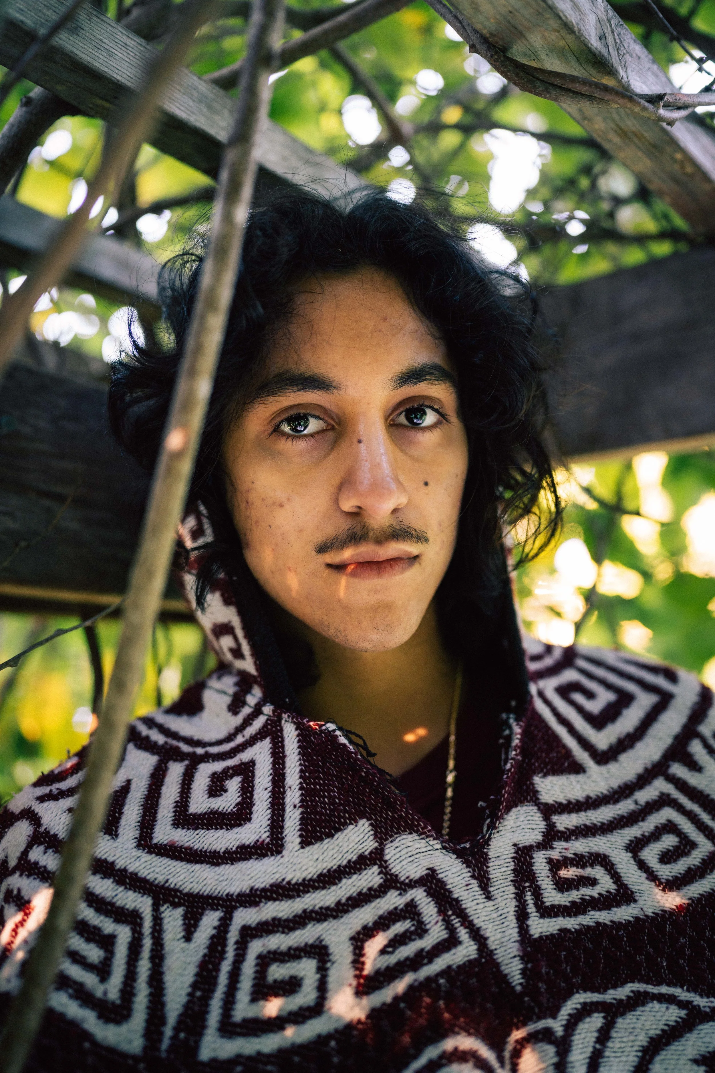 Portrait of a young person with long curly black hair, light brown skin, and facial acne, wearing a patterned hoodie, looking directly at the camera, surrounded by greenery and sunlight filtering through leaves.