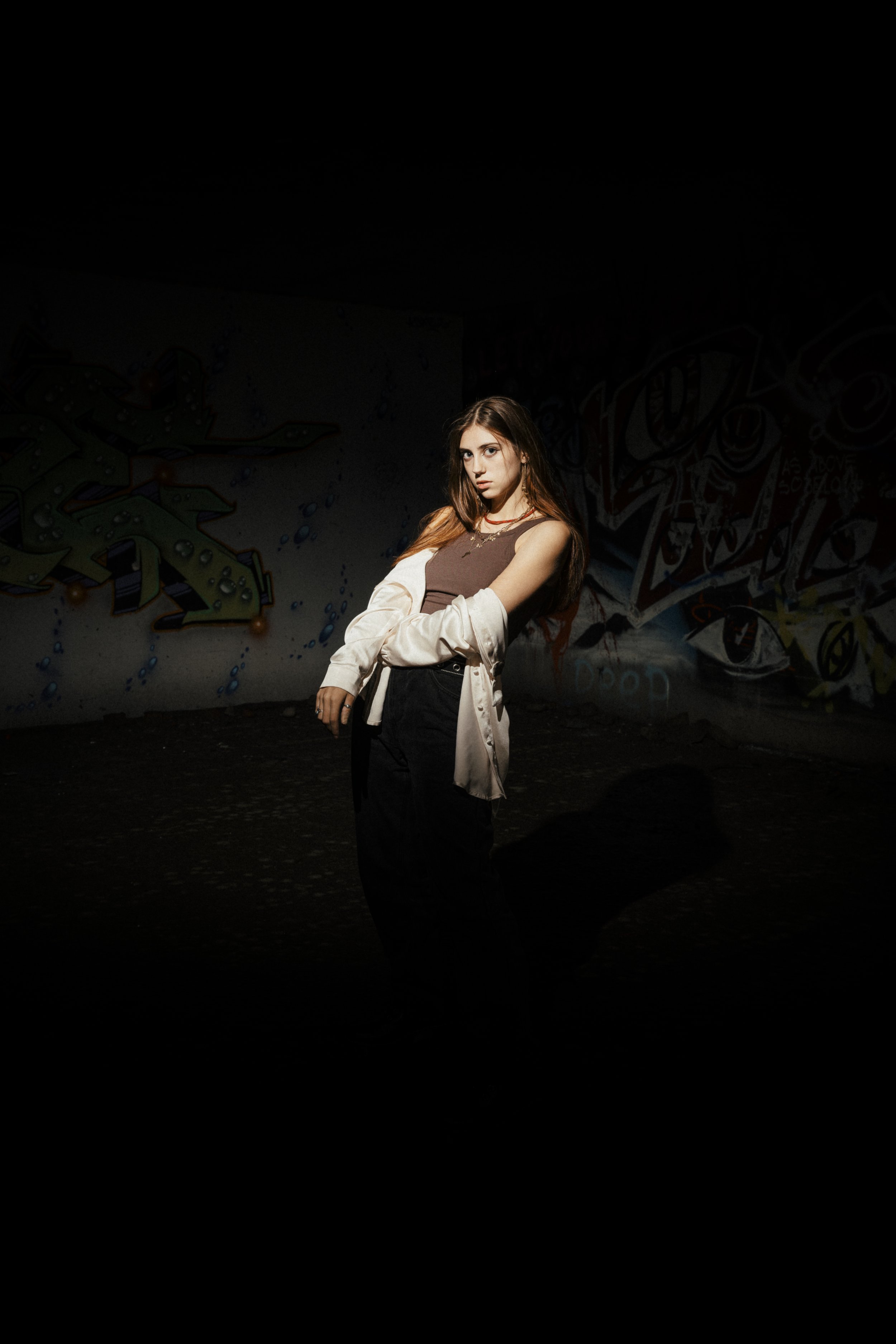 Young woman posing with arms crossed in front of graffiti wall at night with spotlight