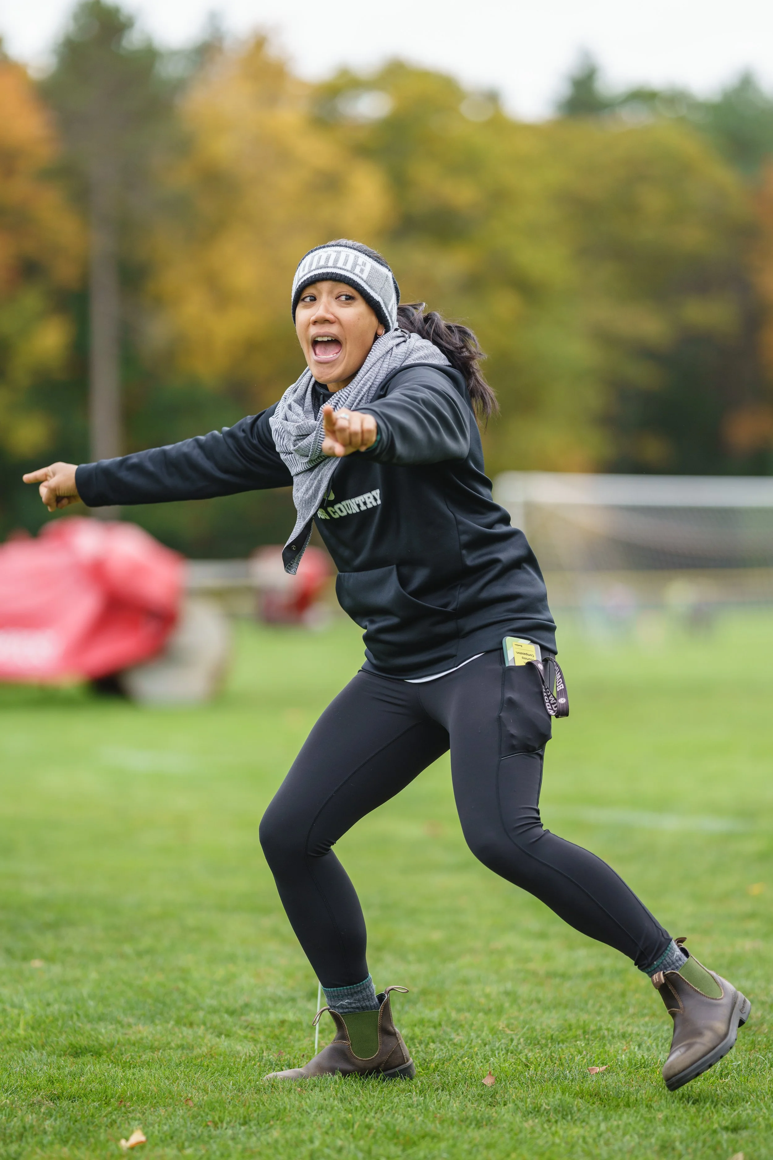 A woman in outdoor sports attire, including a gray beanie, scarf, and black jacket, pointing forward and shouting on a grassy field with a blurred background of trees and a sports goal.