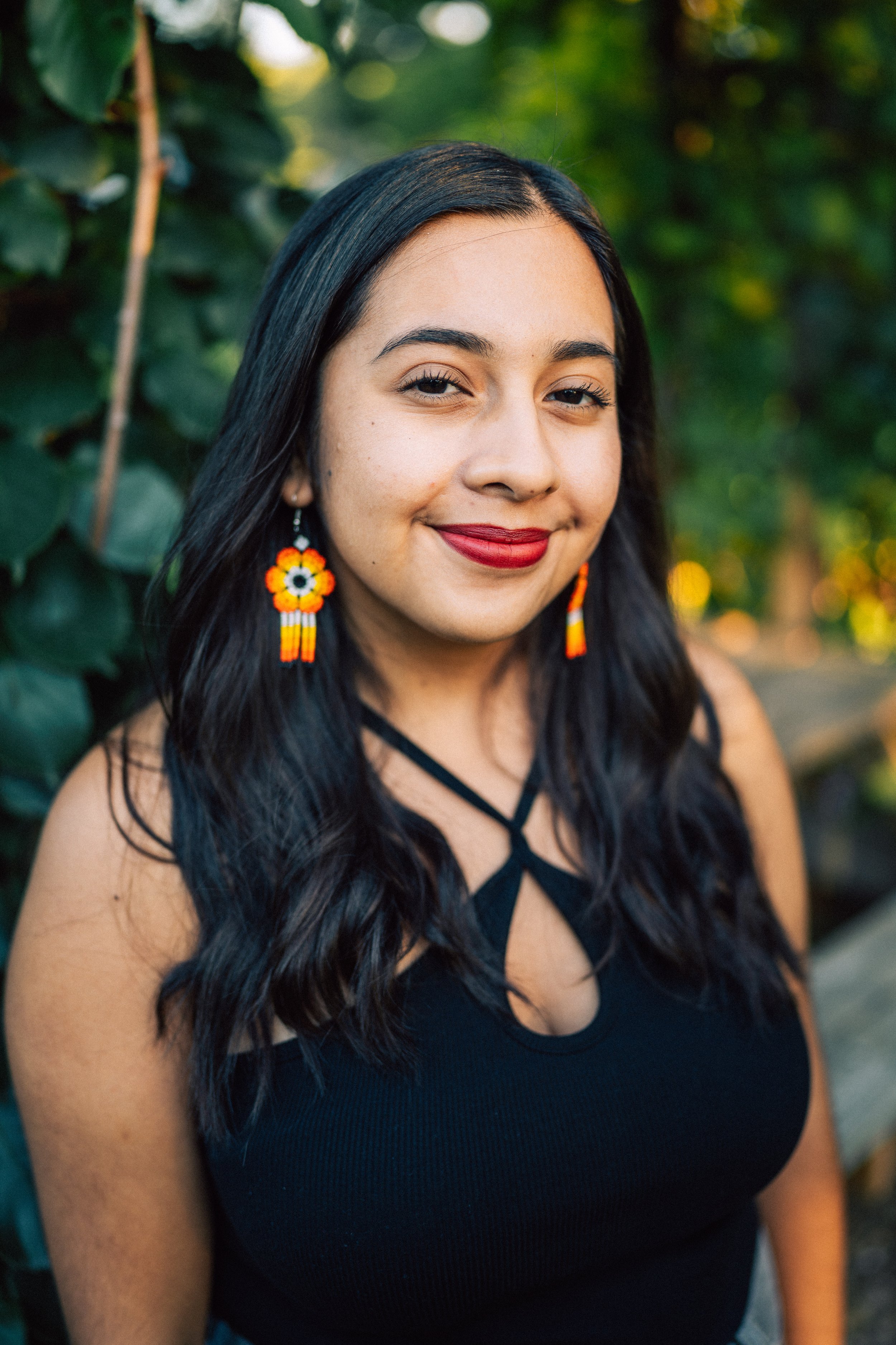 A woman with long dark hair, wearing red lipstick and colorful beaded earrings, smiling outdoors with greenery in the background.