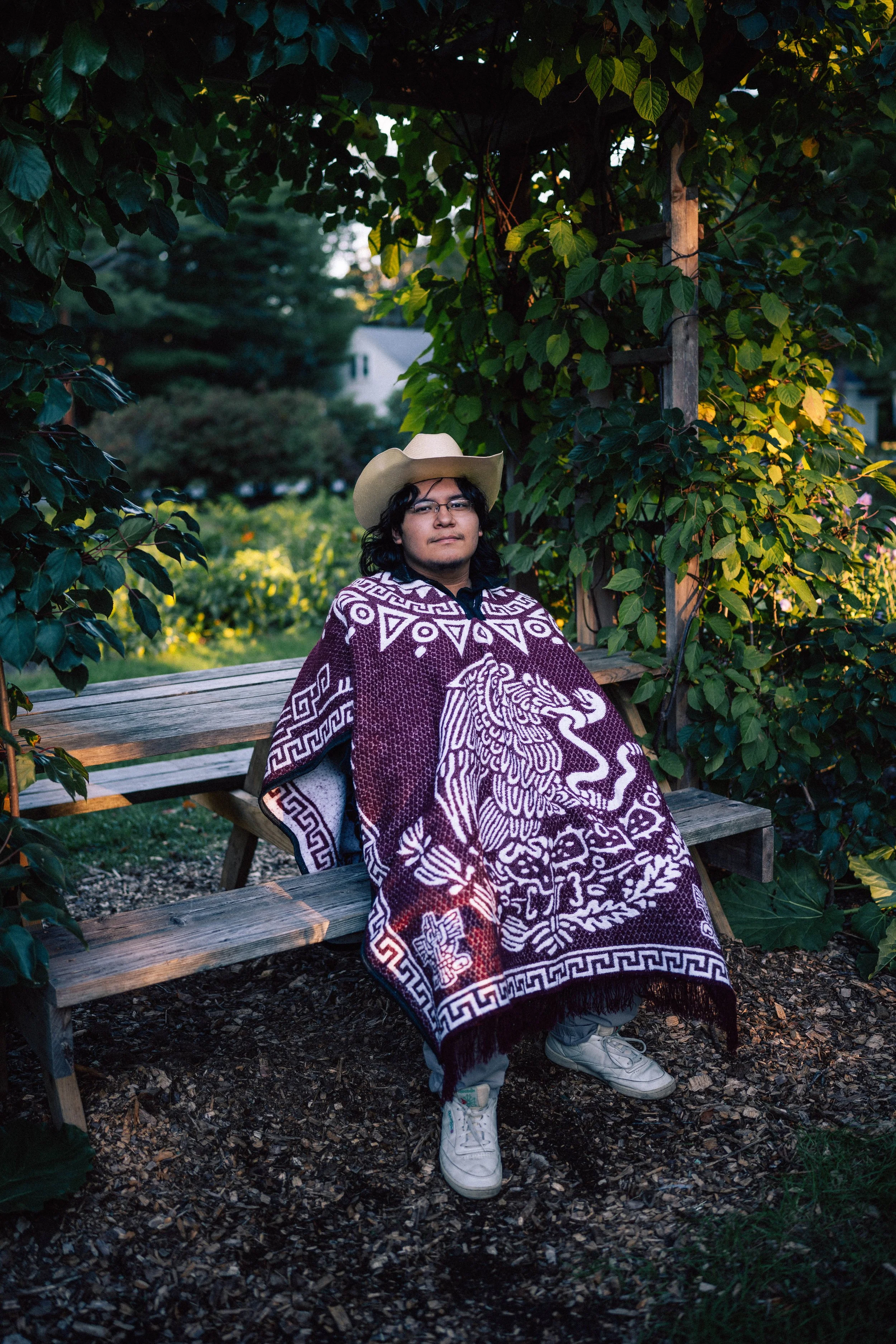 A person wearing glasses, a cowboy hat, and a Mexica design blanket seated on a wooden park bench surrounded by greenery.