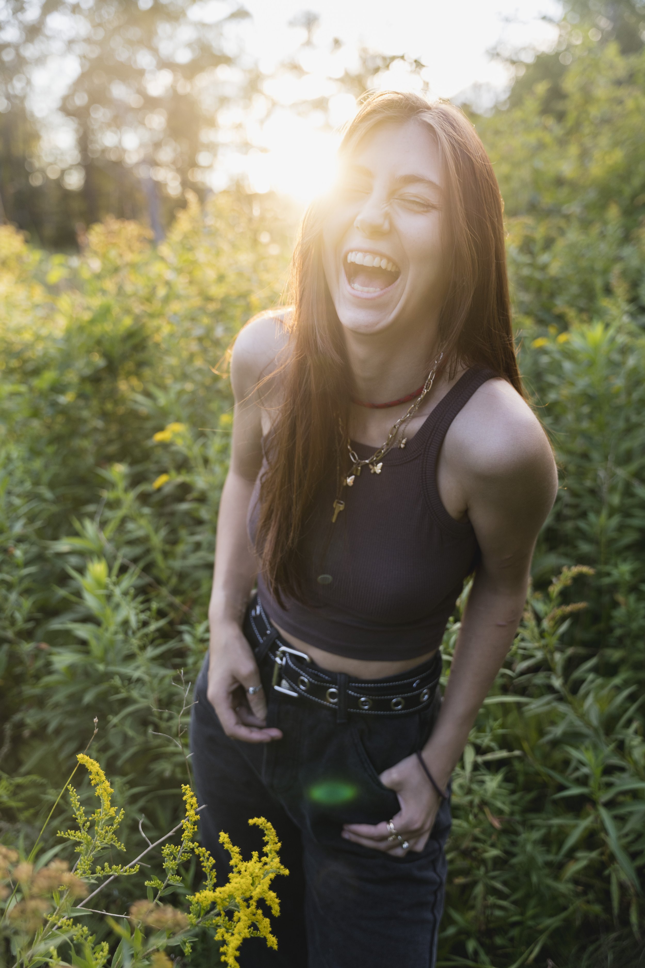 A young woman with long red hair laughing and squinting while standing outdoors in a green, leafy area during sunset.