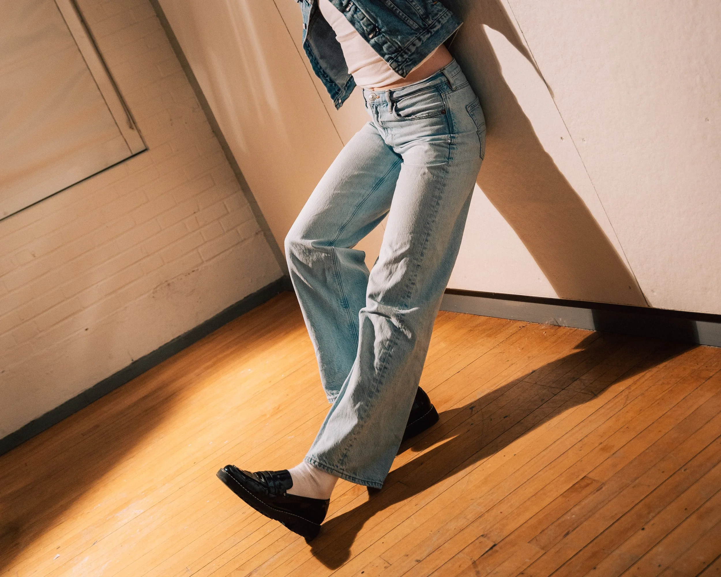 Person leaning against a wall, wearing light blue jeans, a denim jacket, and black shoes, standing on a wooden floor.