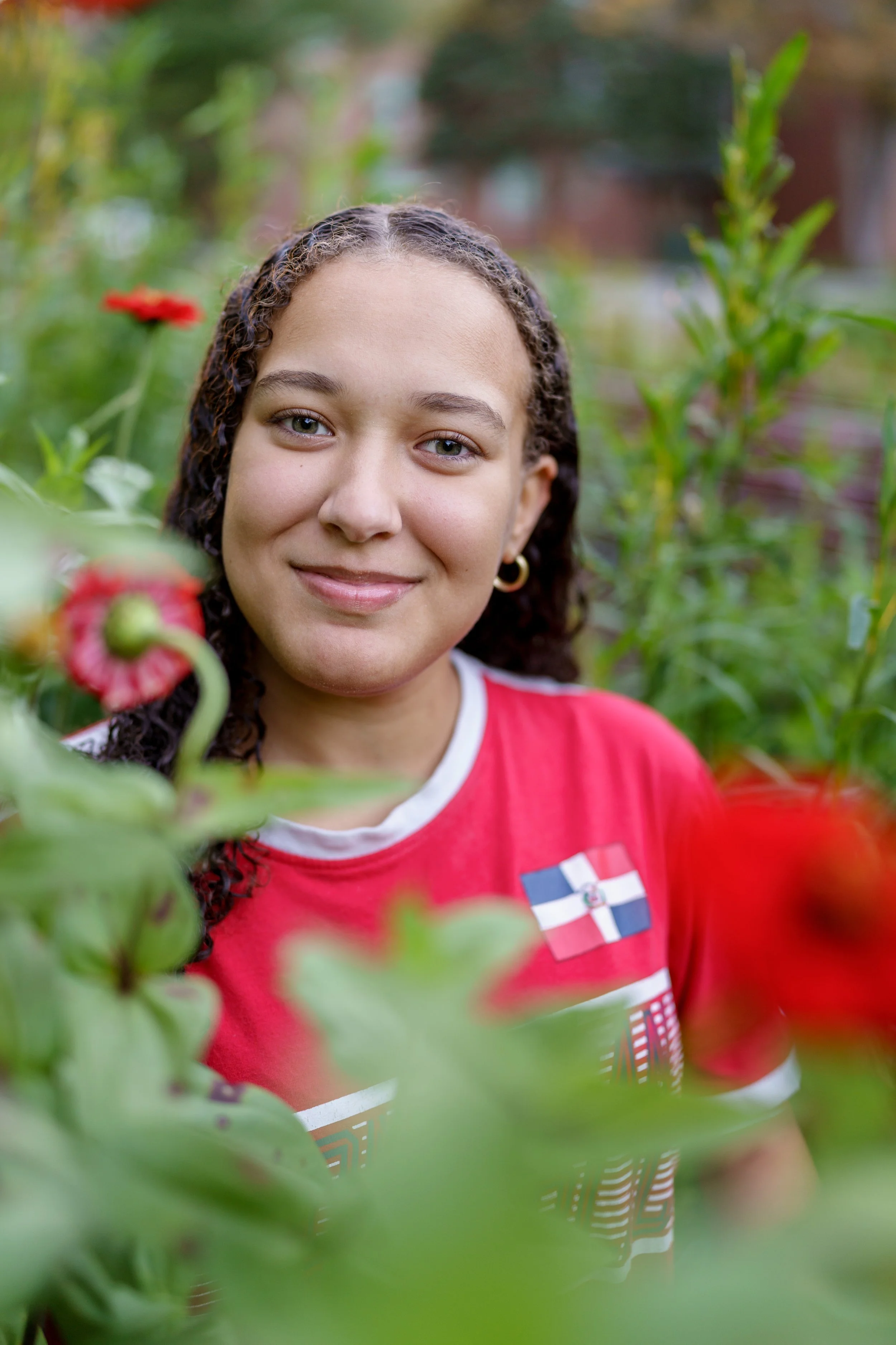 A young woman with curly hair, wearing a red shirt with the Dominican Republic flag, smiles while standing in a garden surrounded by green plants and red flowers.