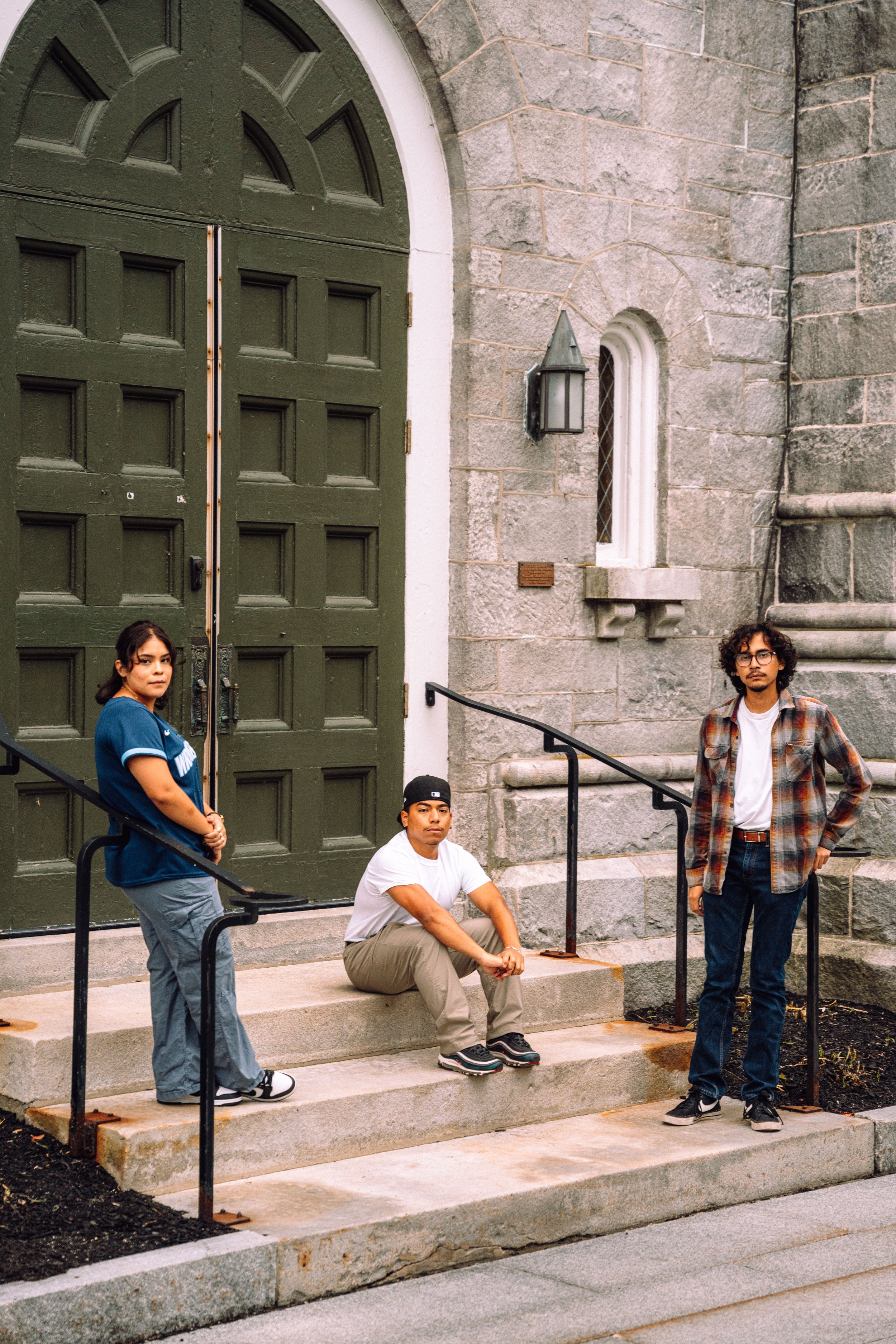 Three young adults posing on stone steps in front of a large green wooden door and stone building.
