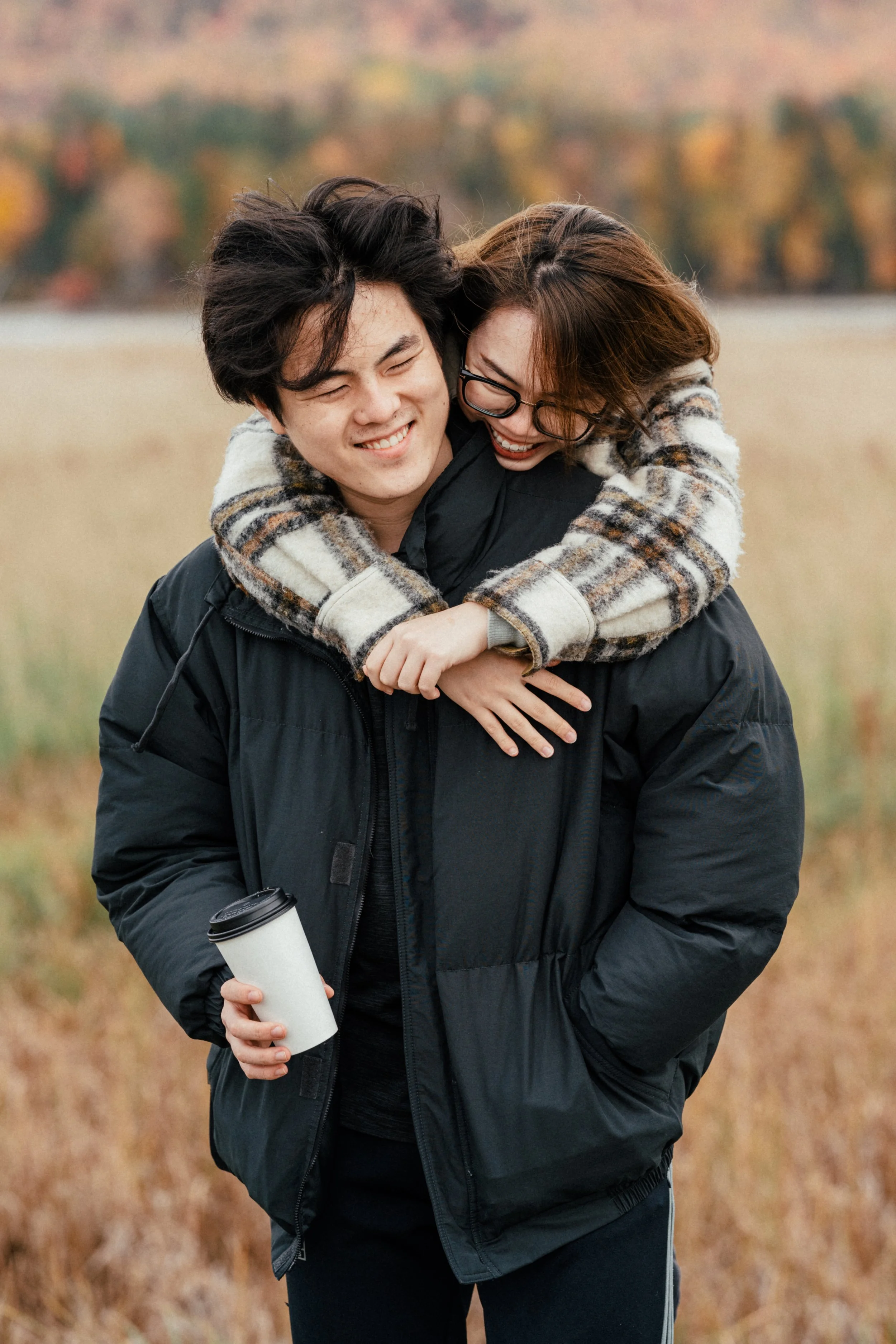 A young couple smiling and laughing outdoors, the woman hugging the man from behind, holding onto his shoulders, with a background of fall foliage.