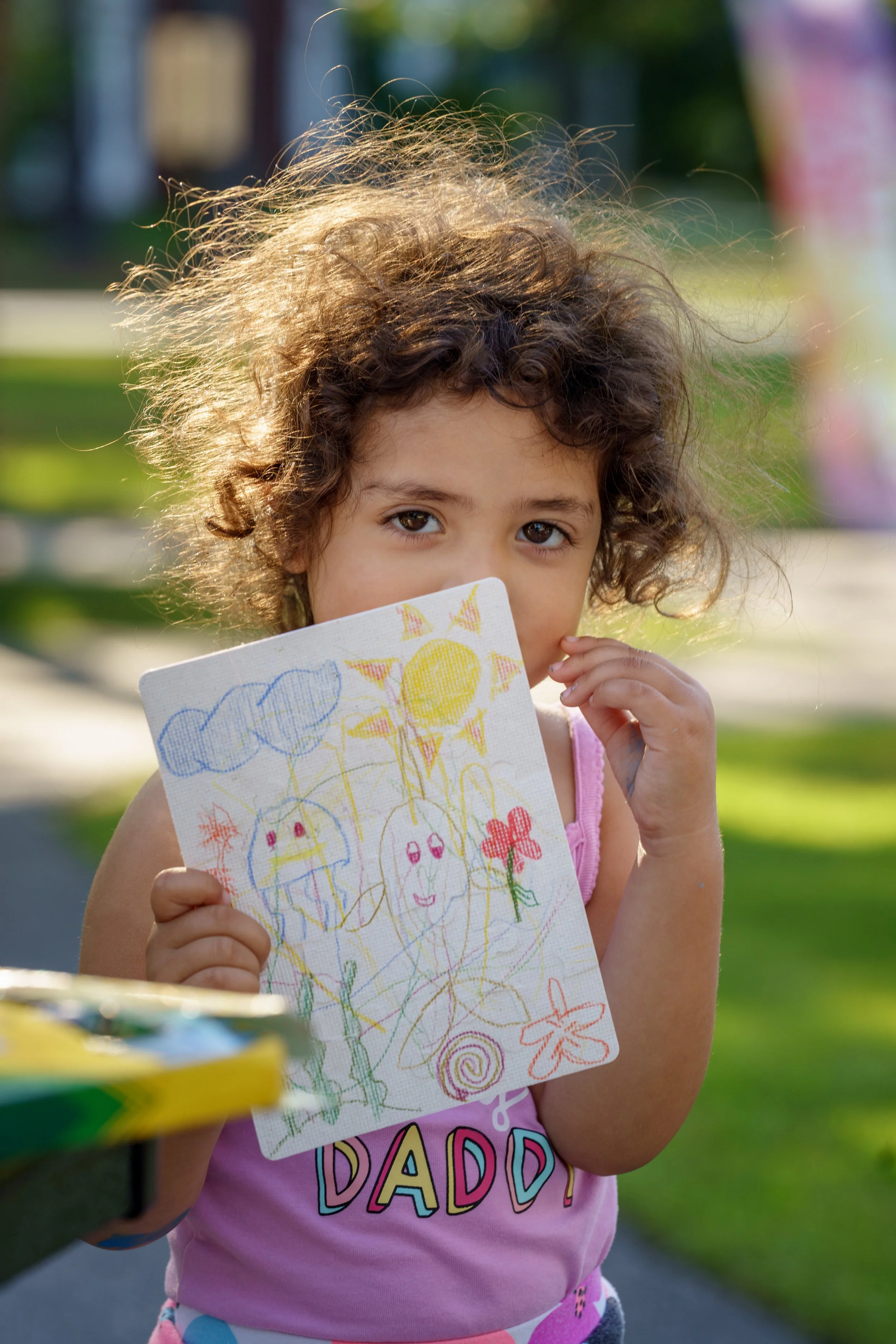 A young girl with curly hair holding a colorful drawing of smiling suns, clouds, flowers, and swirls, standing outdoors on a sunny day.