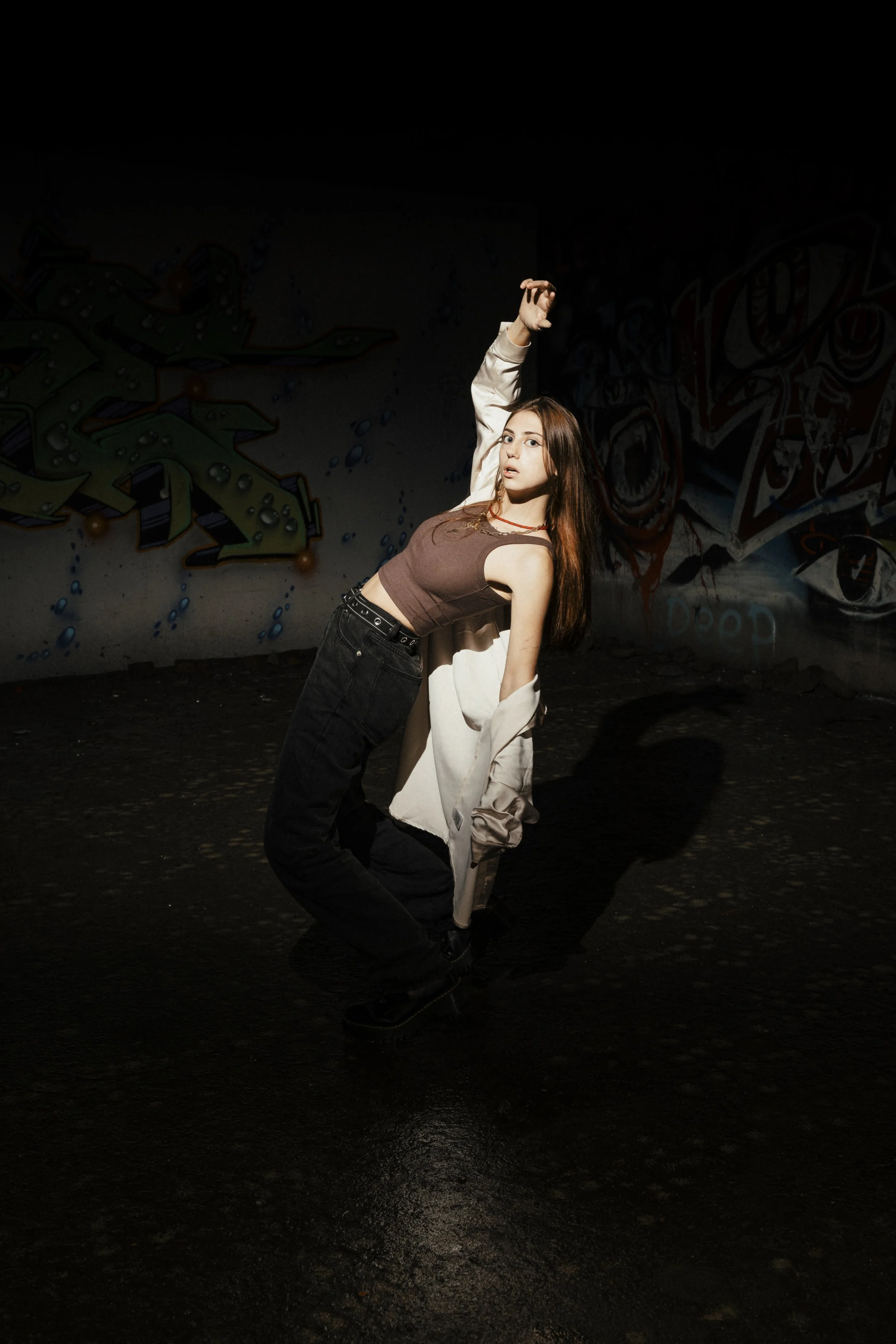 A young woman dancing in a dimly lit space with graffiti on the wall behind her.