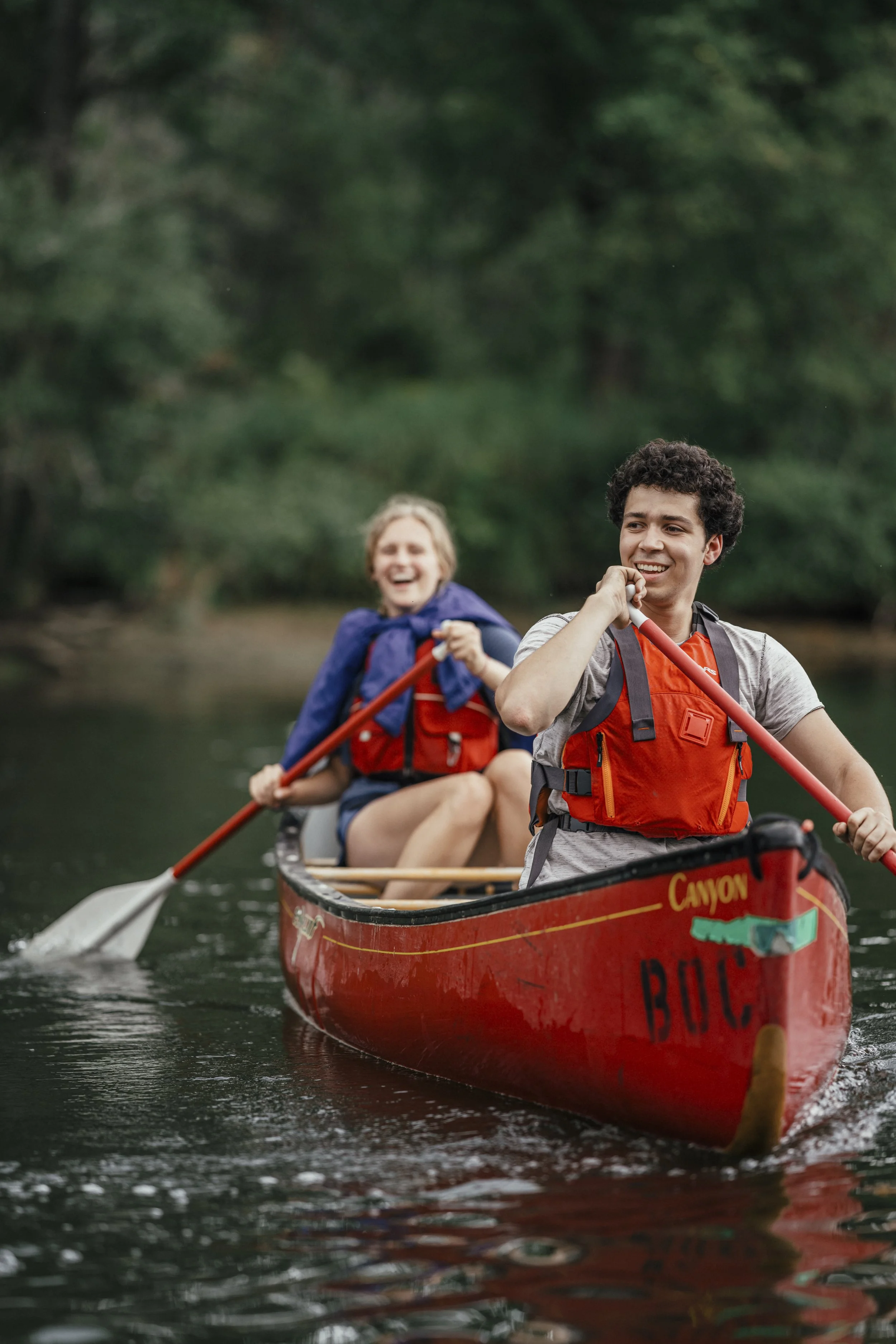Two people enjoying a canoe ride on a river surrounded by trees, both wearing life jackets, smiling and paddling.