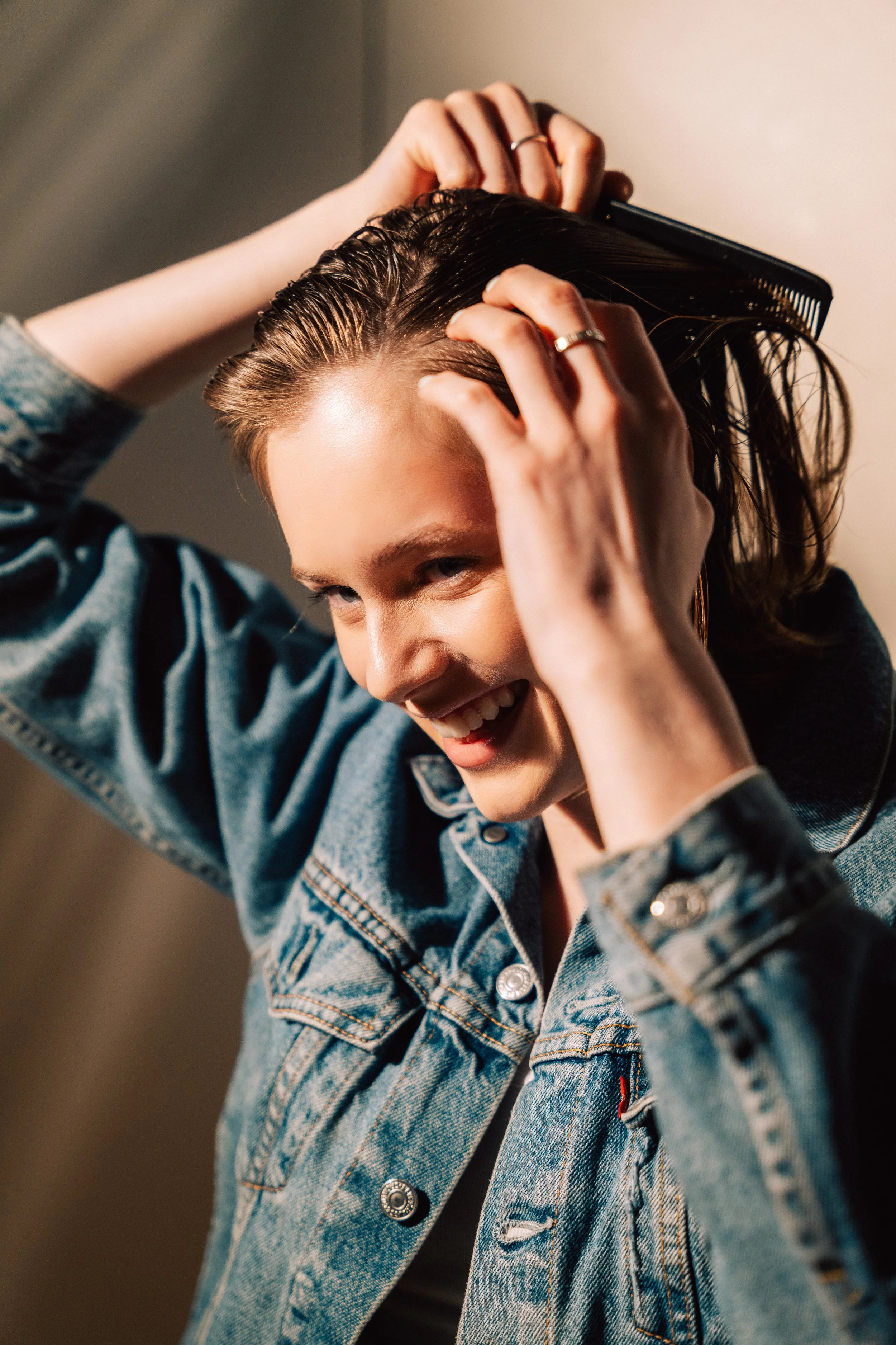 Woman with wet hair combing it back, smiling in a denim jacket.