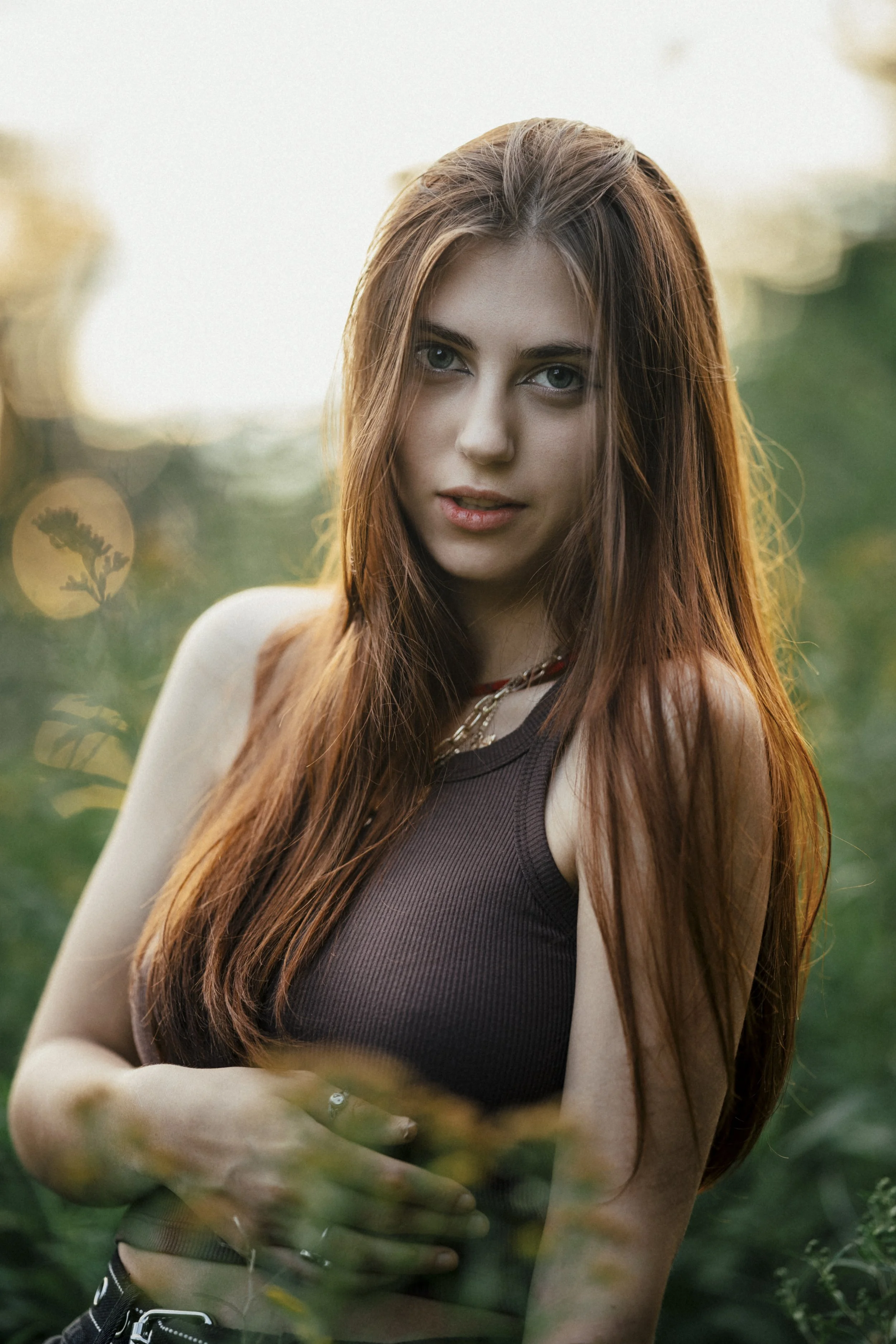 A young woman with long red hair and green eyes standing outdoors in a natural setting with soft sunlight, looking at the camera.