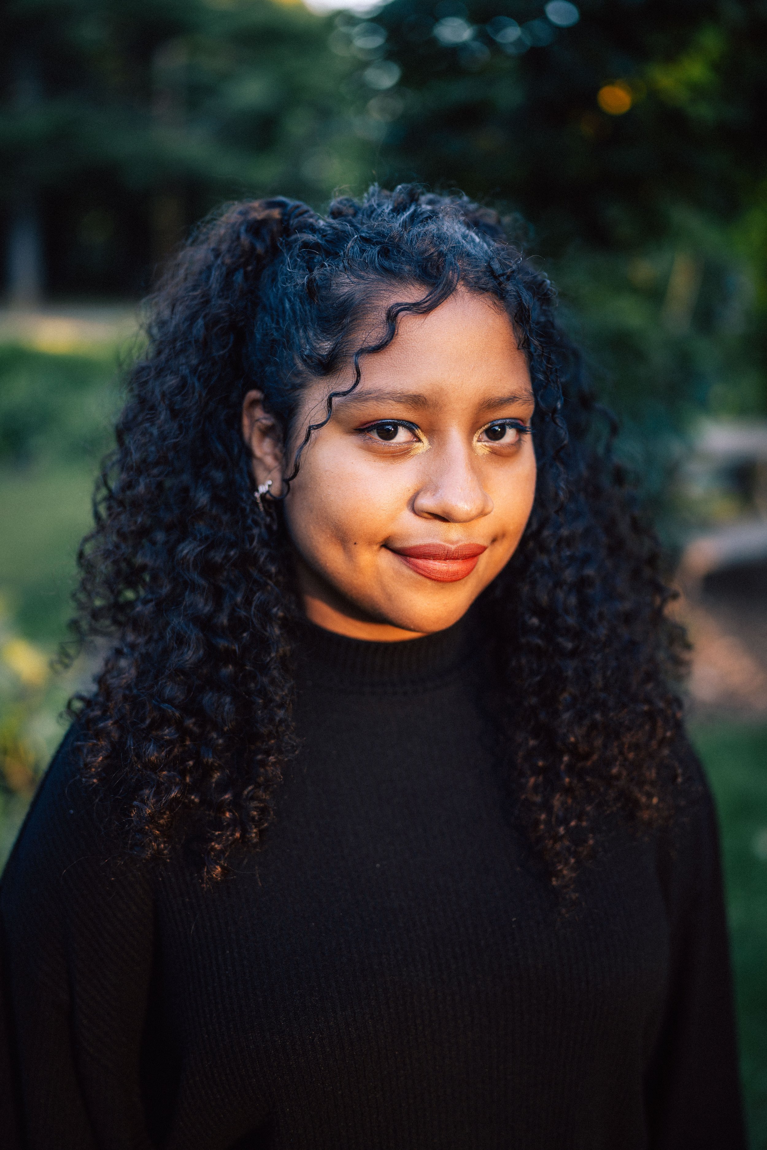 A young woman with dark, curly hair and tan skin smiling outdoors at sunset, wearing a black top and gold eyeshadow.