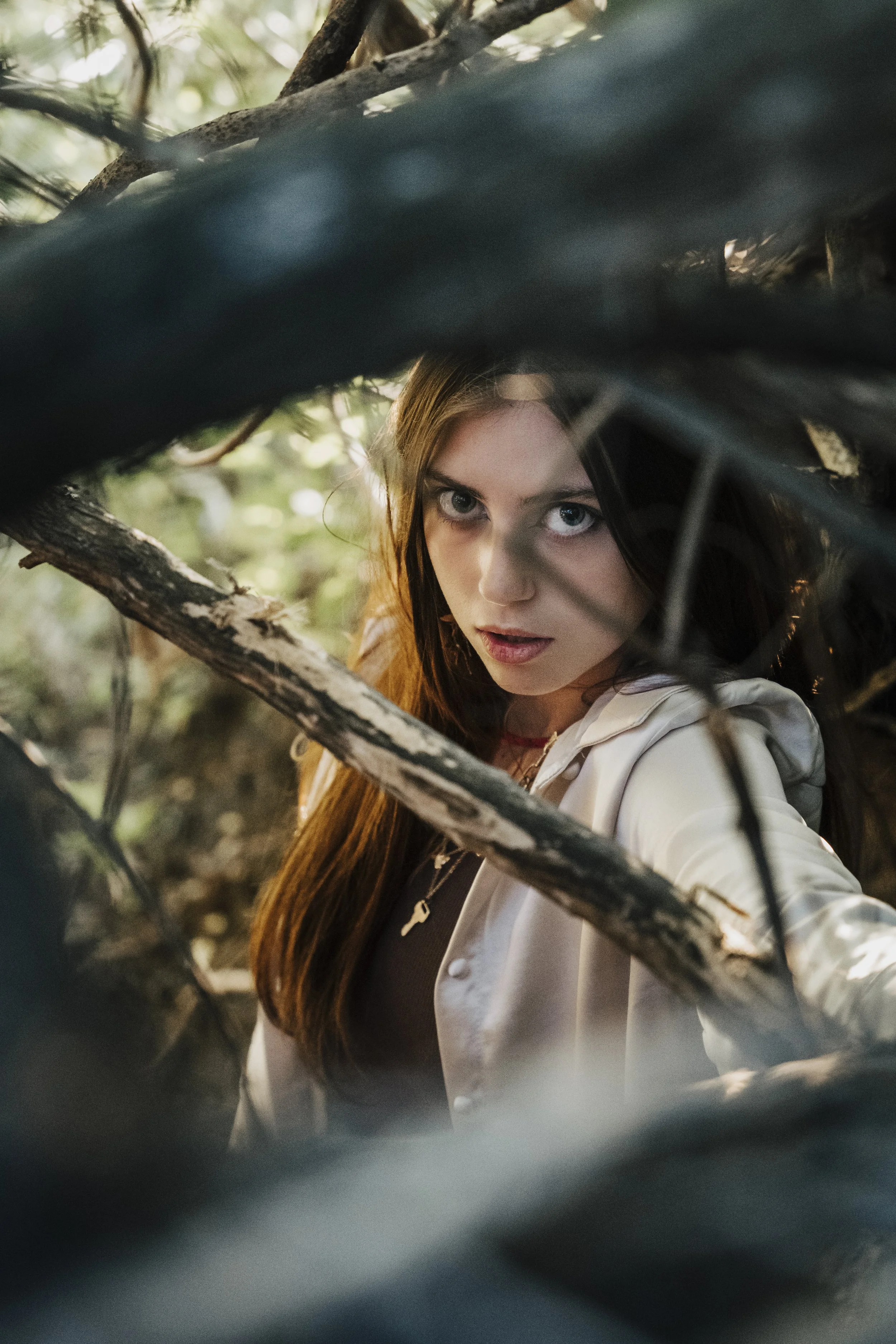 A young woman with long brown hair and blue eyes looking through tree branches in a forest.