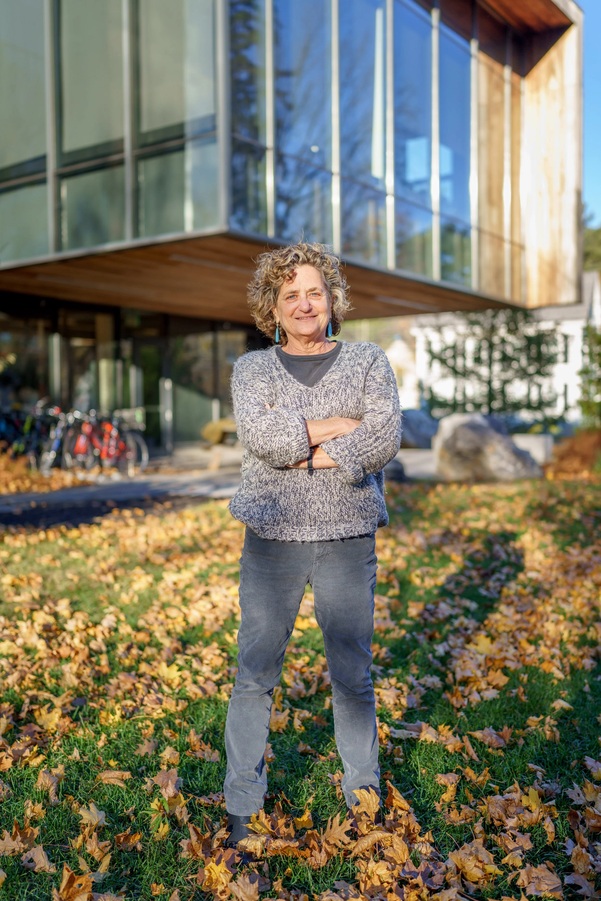 A woman with curly gray hair standing on a grassy area covered with fallen autumn leaves, smiling with arms crossed, in front of a modern building with large glass windows and a wooden overhang.