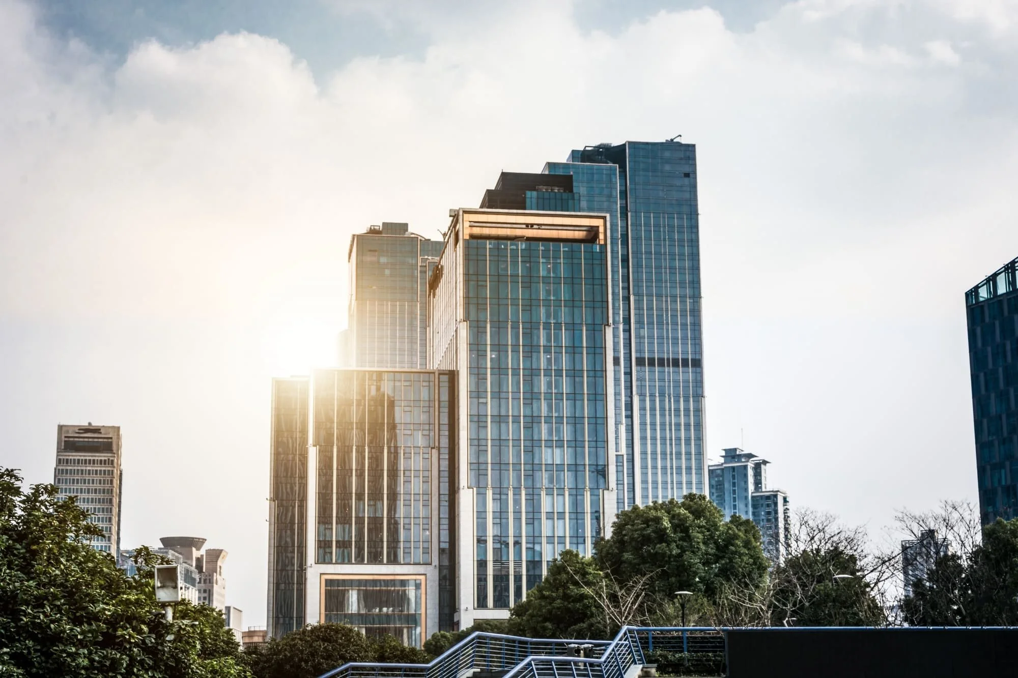 Modern glass skyscrapers under a partly cloudy sky with sunlight reflecting off the buildings.