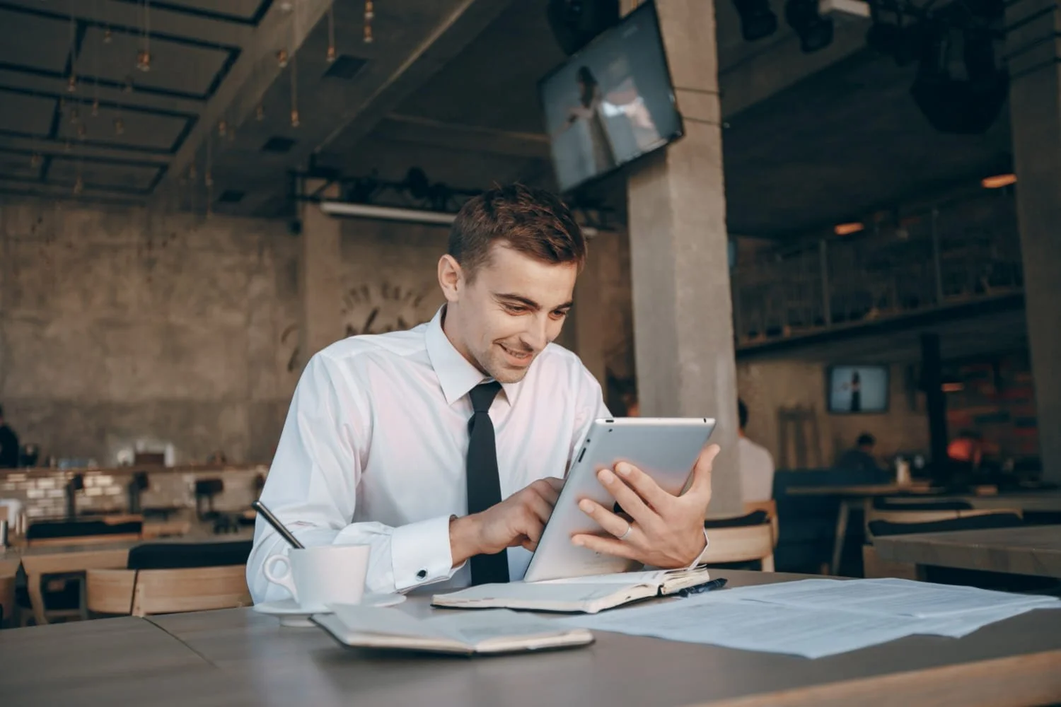 A young man in a white dress shirt and black tie sitting at a table in a modern cafe, smiling as he interacts with a tablet. The table has newspapers, a coffee mug, and a pen. There are multiple TVs and other people in the background.