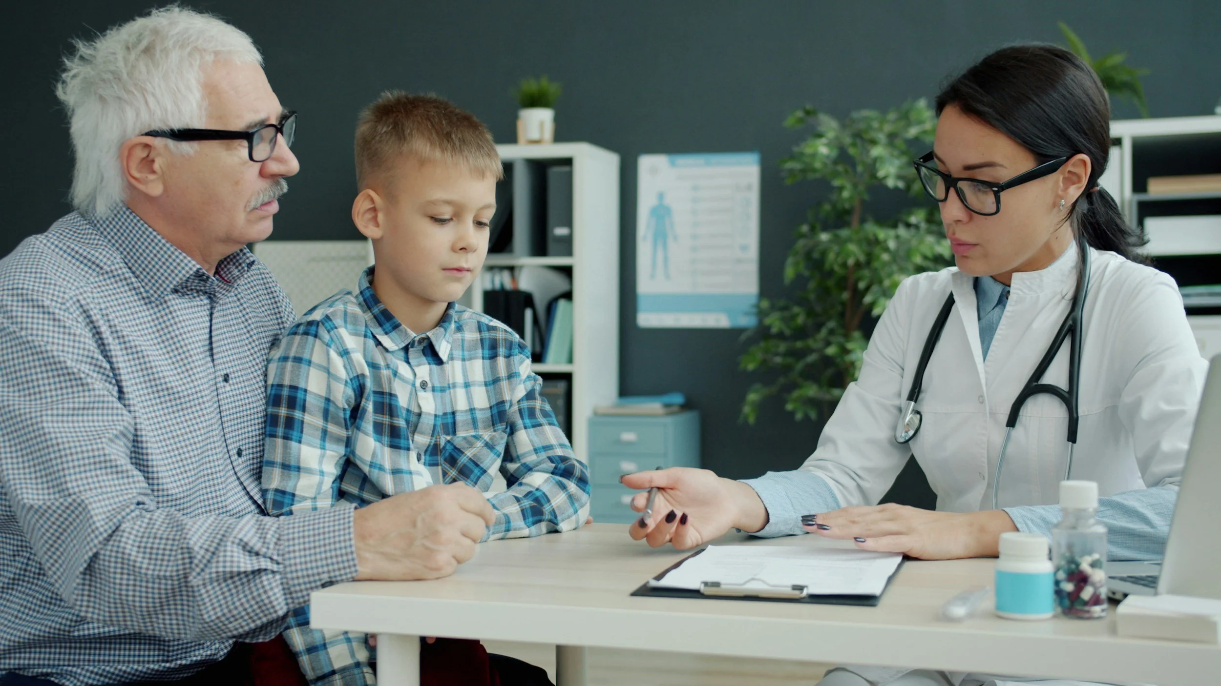 A young boy sitting at a doctor's office with an older man, possibly his grandfather, while a female doctor examines him. The girl doctor has a stethoscope around her neck and a clipboard on the table.