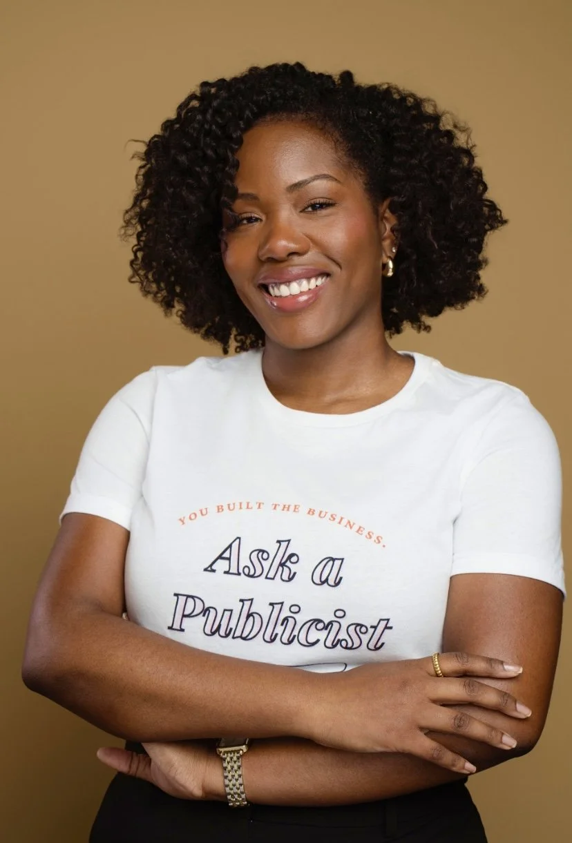A smiling Black woman with curly hair, wearing a white t-shirt that says 'Ask a Publicist' and a gold watch, standing against a plain beige background.