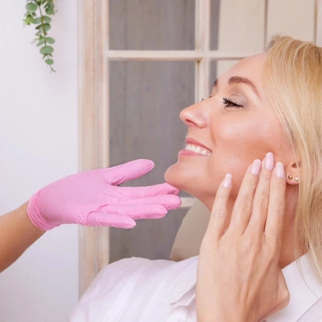 A woman with blonde hair smiling with closed eyes, touching her face, while a person in pink gloves gently holds her chin in a skincare or beauty treatment setting.